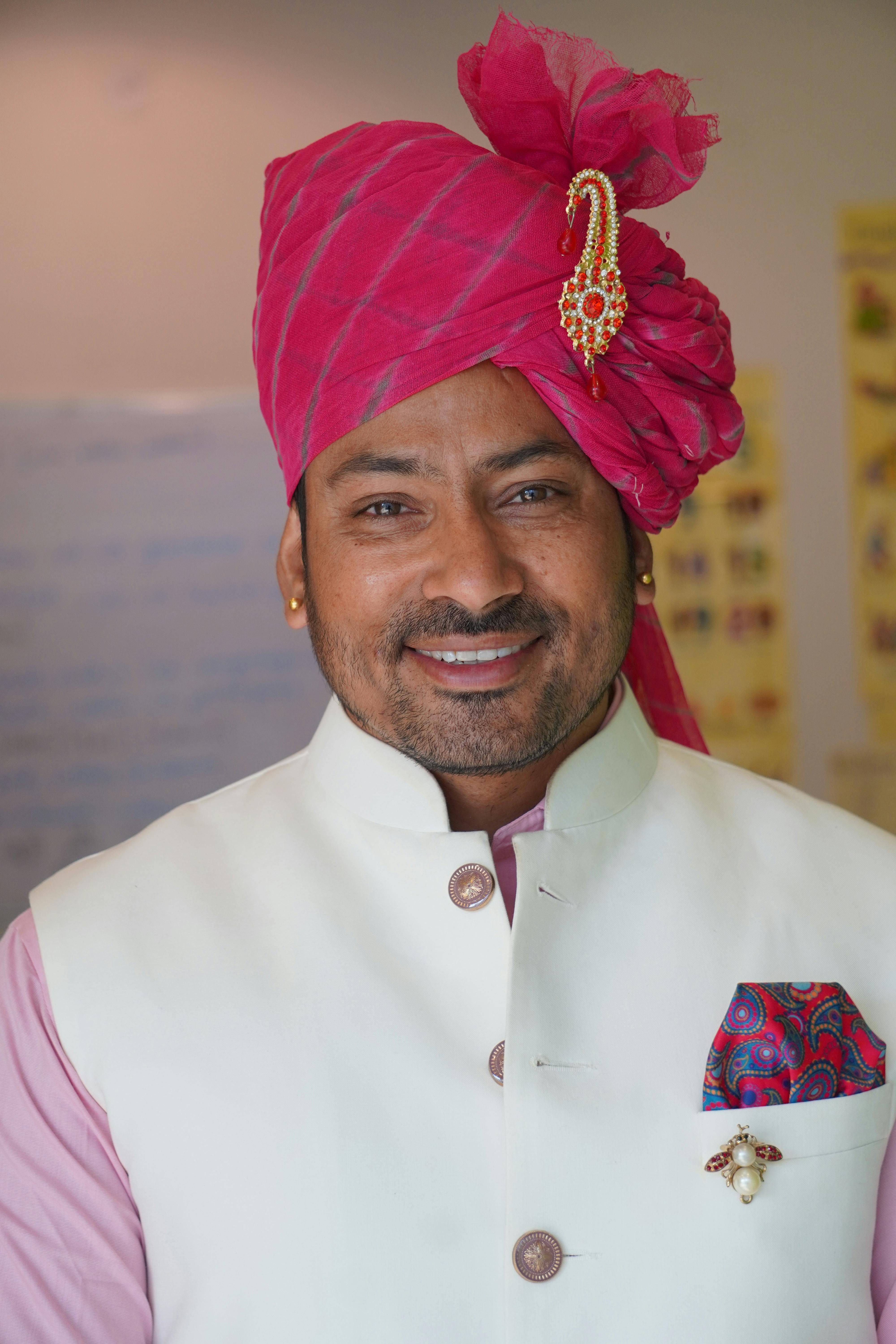 A man wearing traditional Indian turban and jacket smiles warmly indoors.