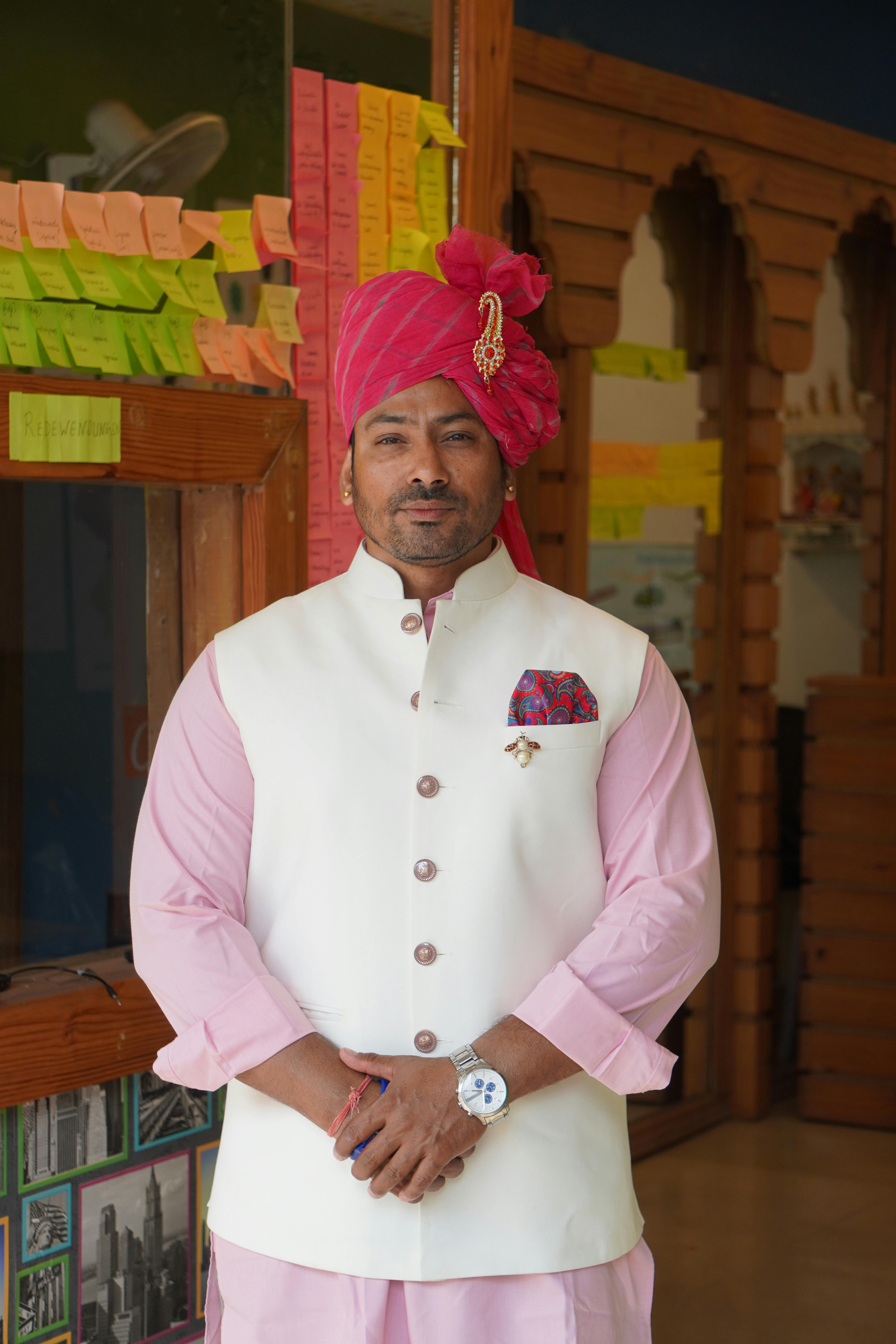 A man in pink traditional Indian attire poses indoors with colorful notes background.