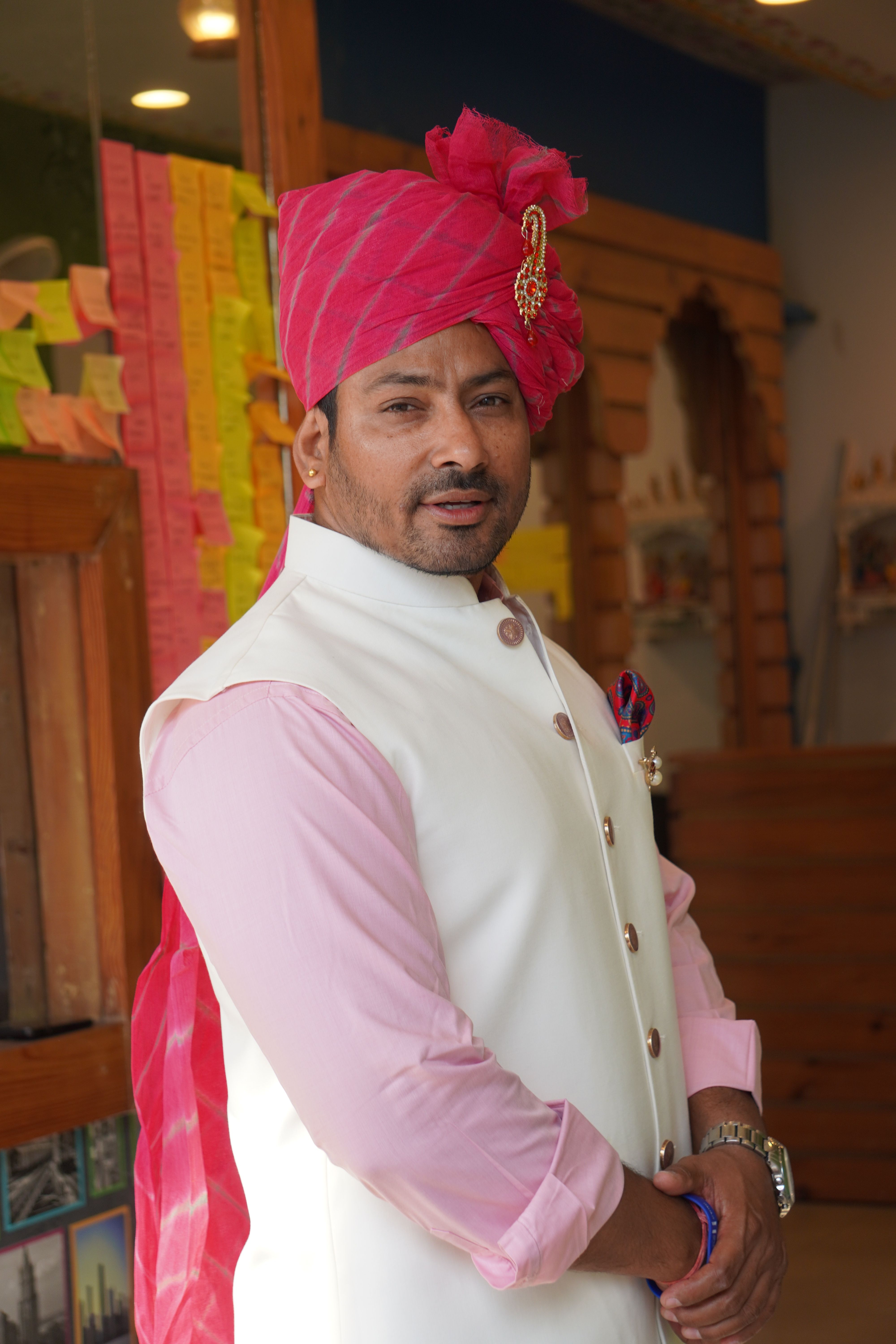 A man wearing traditional Indian wedding attire, including a pink turban, posing indoors.