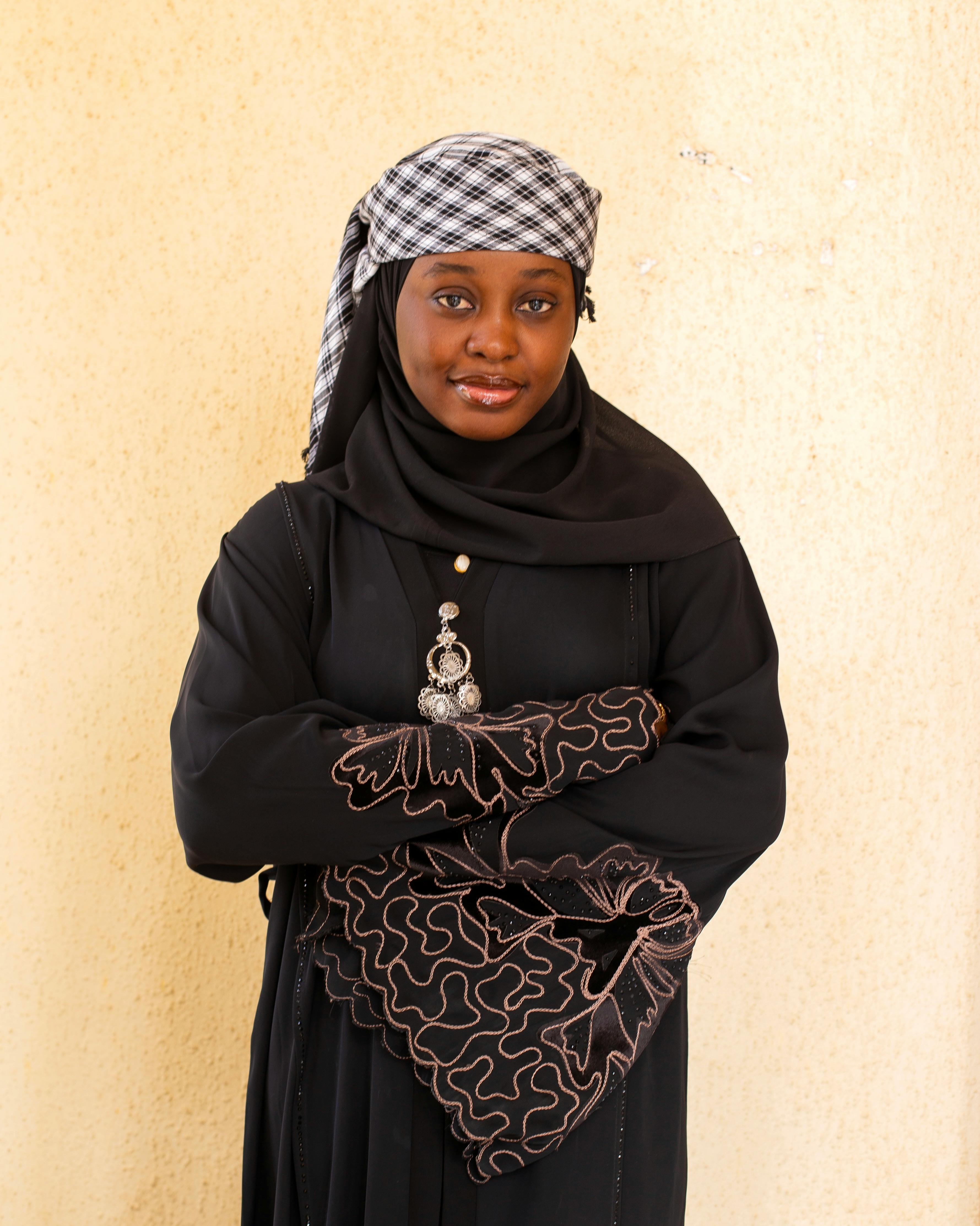 Portrait of a confident woman in a black hijab with intricate embroidery against a neutral background.