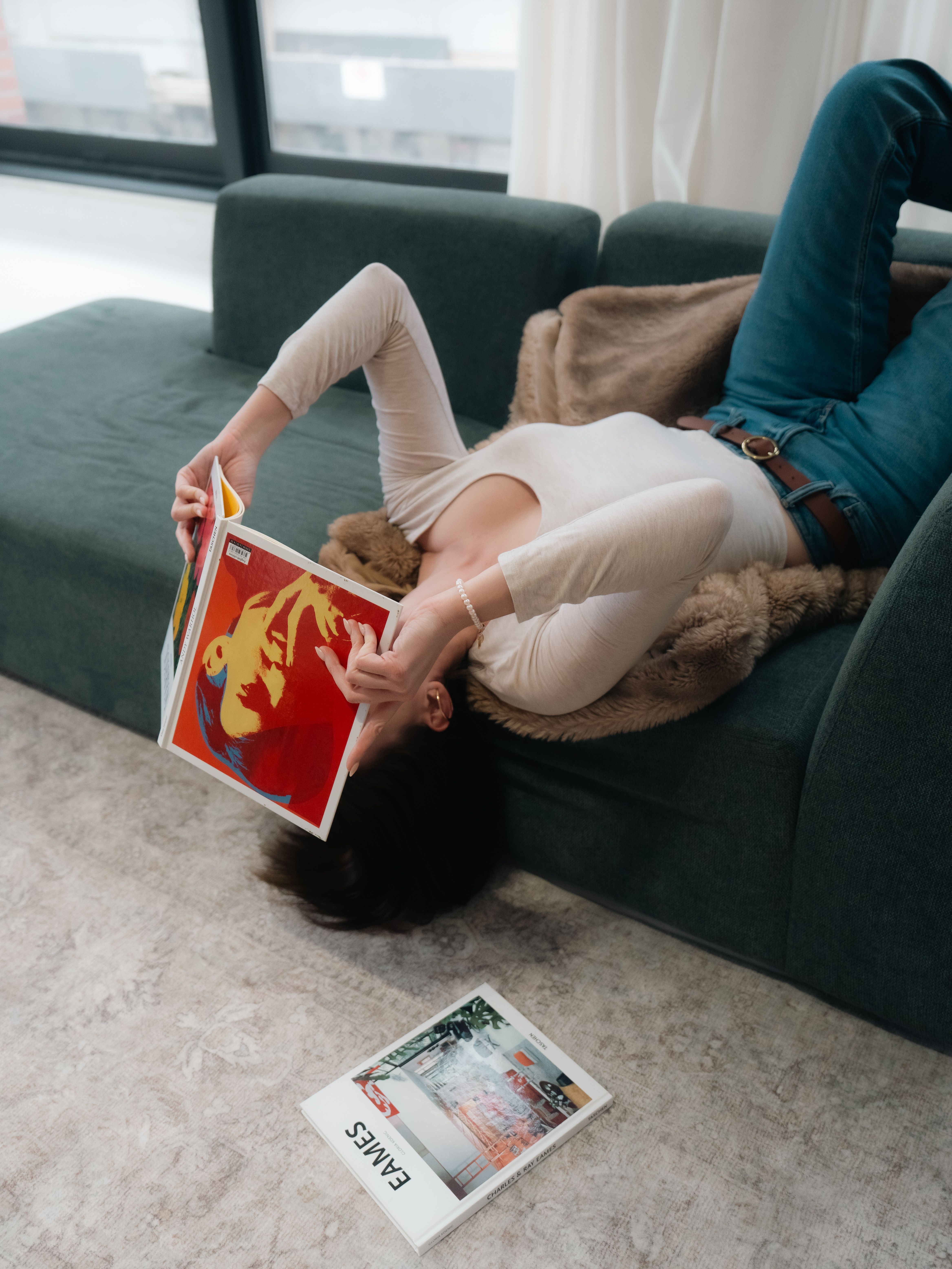 Casual scene of a woman lying on a sofa reading magazines with natural light streaming in.