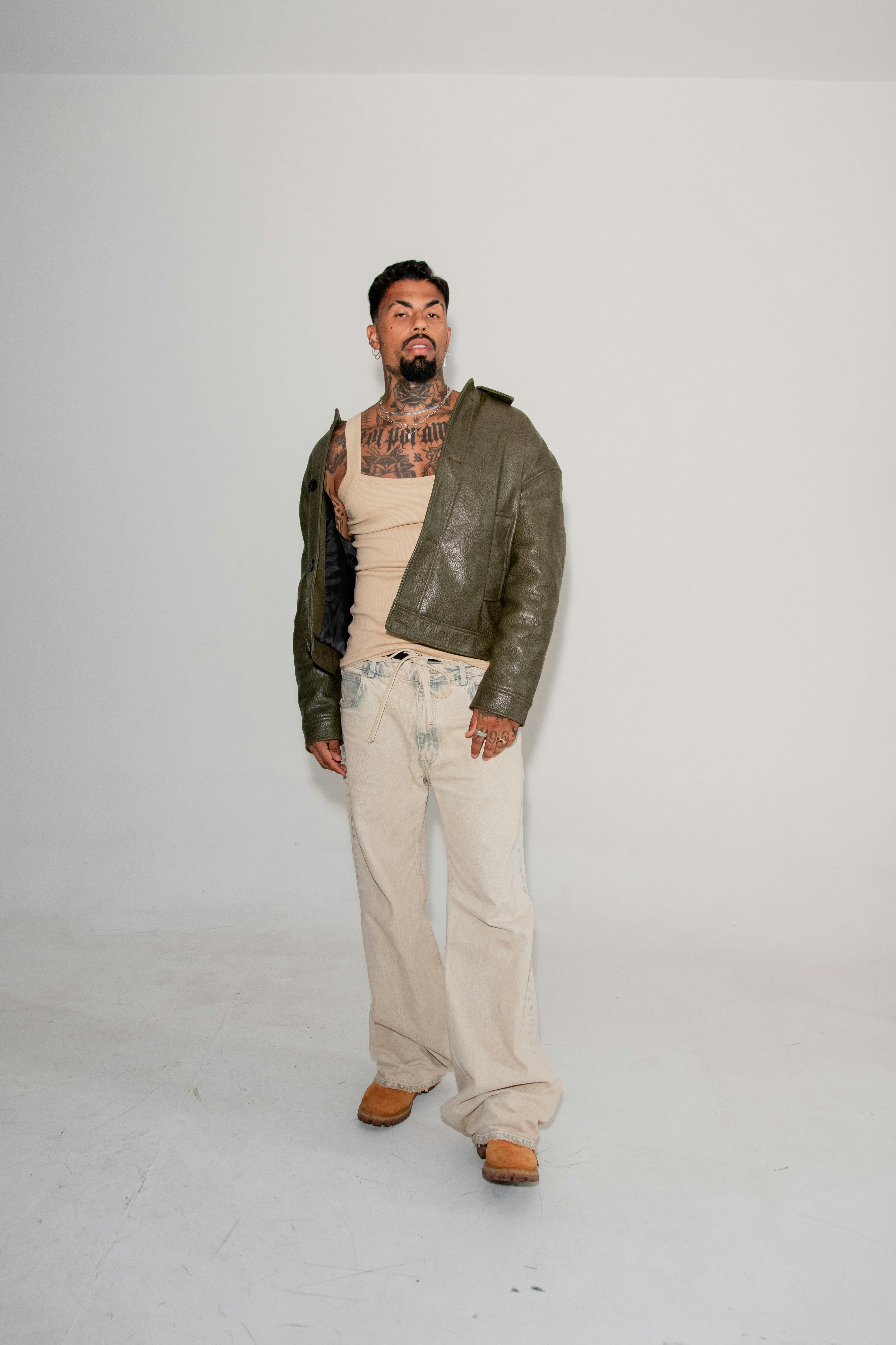 무료 녹색 가죽 재킷과 베이지색 바지를 입은 세련된 남성이 밝은 배경 앞에서 자신감 넘치는 포즈를 취하고 있다. 스톡 사진