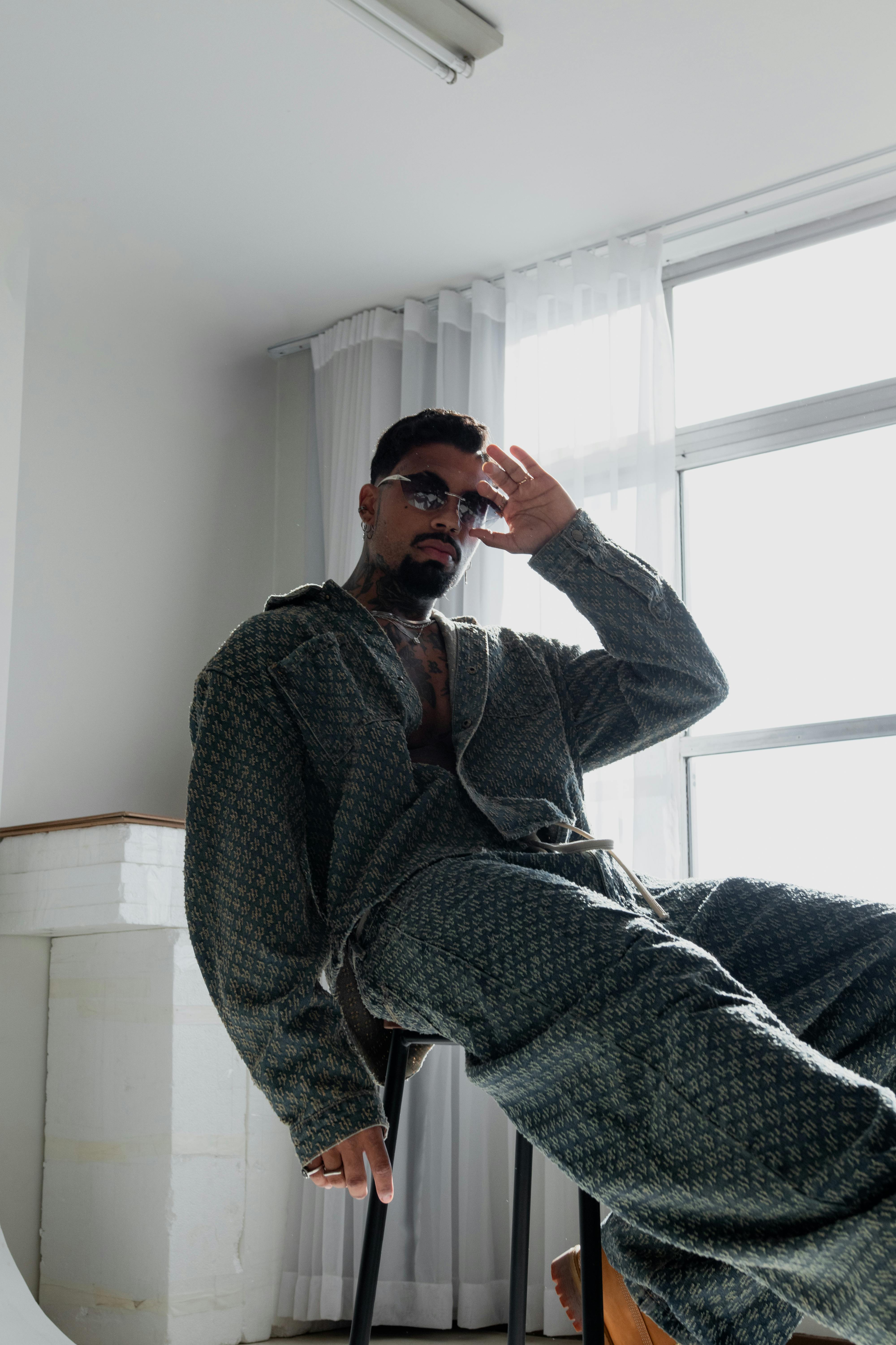 Free A fashionable man in a patterned outfit lounges by a bright window, creating a modern indoor look. Stock Photo