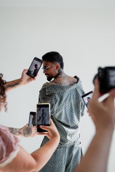 A man with tattoos poses while multiple people capture his image using smartphones.