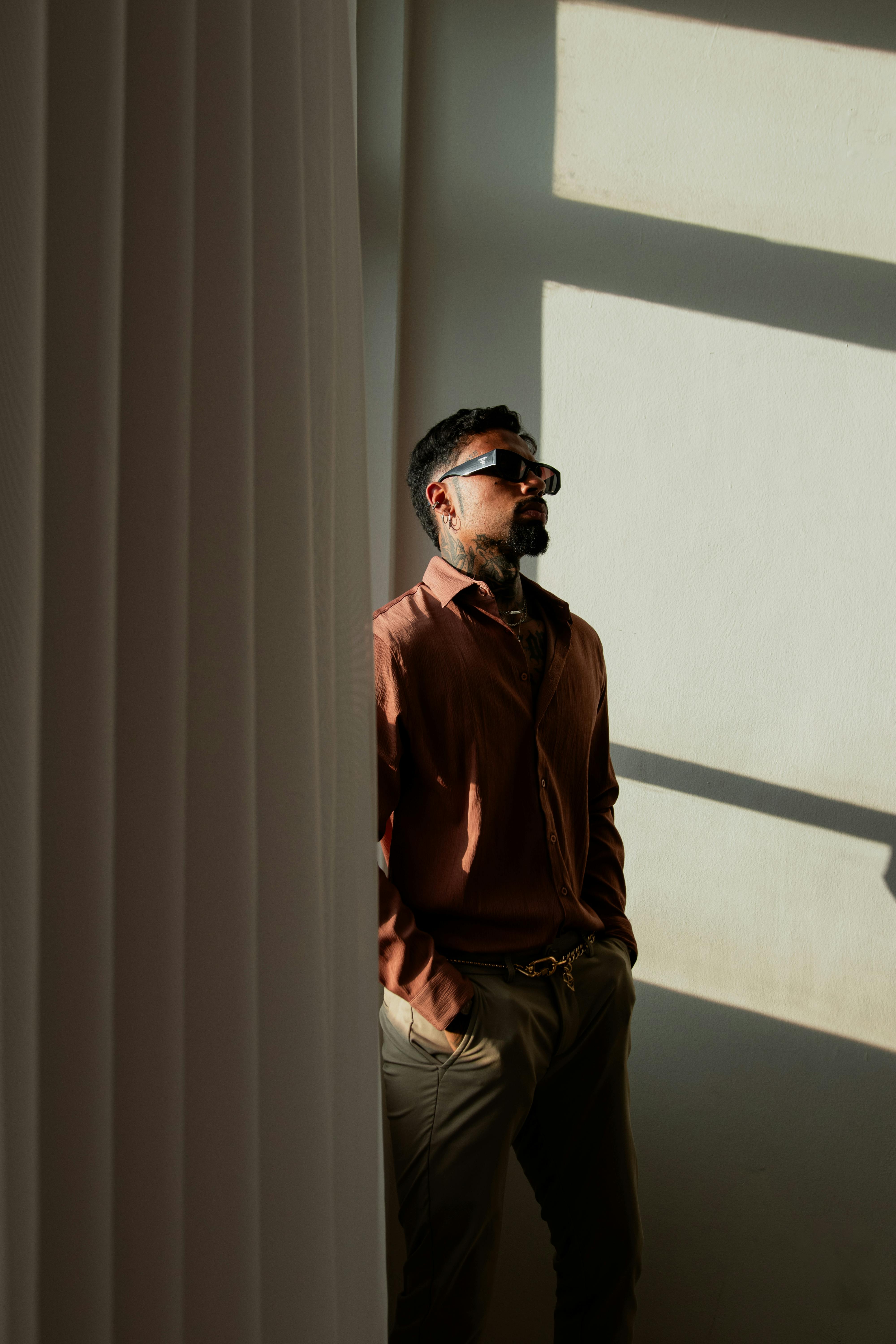 Free A fashionable man in sunglasses stands against a sunlit wall, exuding style and confidence. Stock Photo