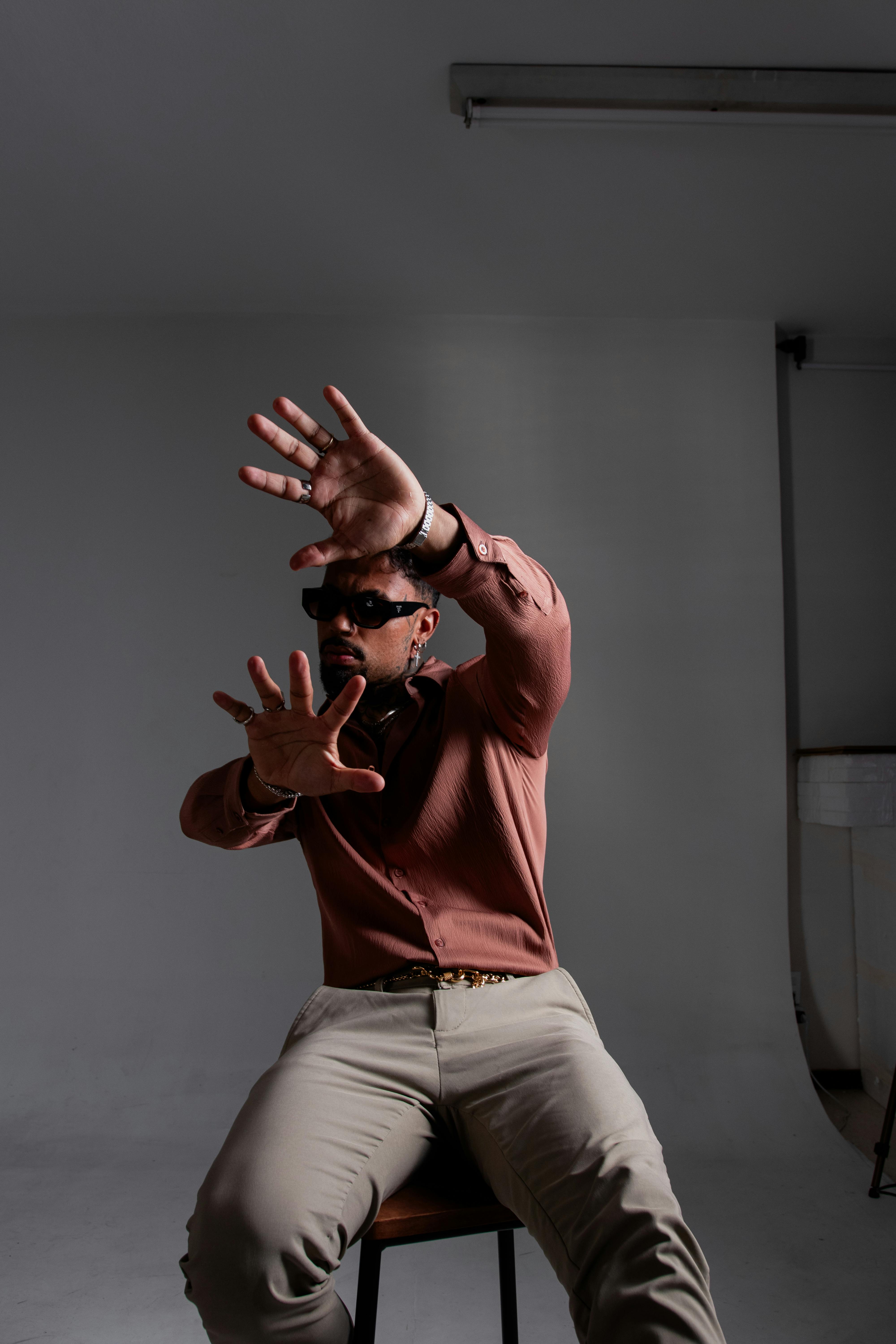 無料 サングラスをかけ、ボタン付きのシャツを着て、屋内に座り、手を上げて身振りをしている男性。 写真素材