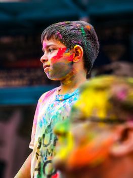 A young boy with painted face stands during Holi festival, vibrant colors draping his clothes.