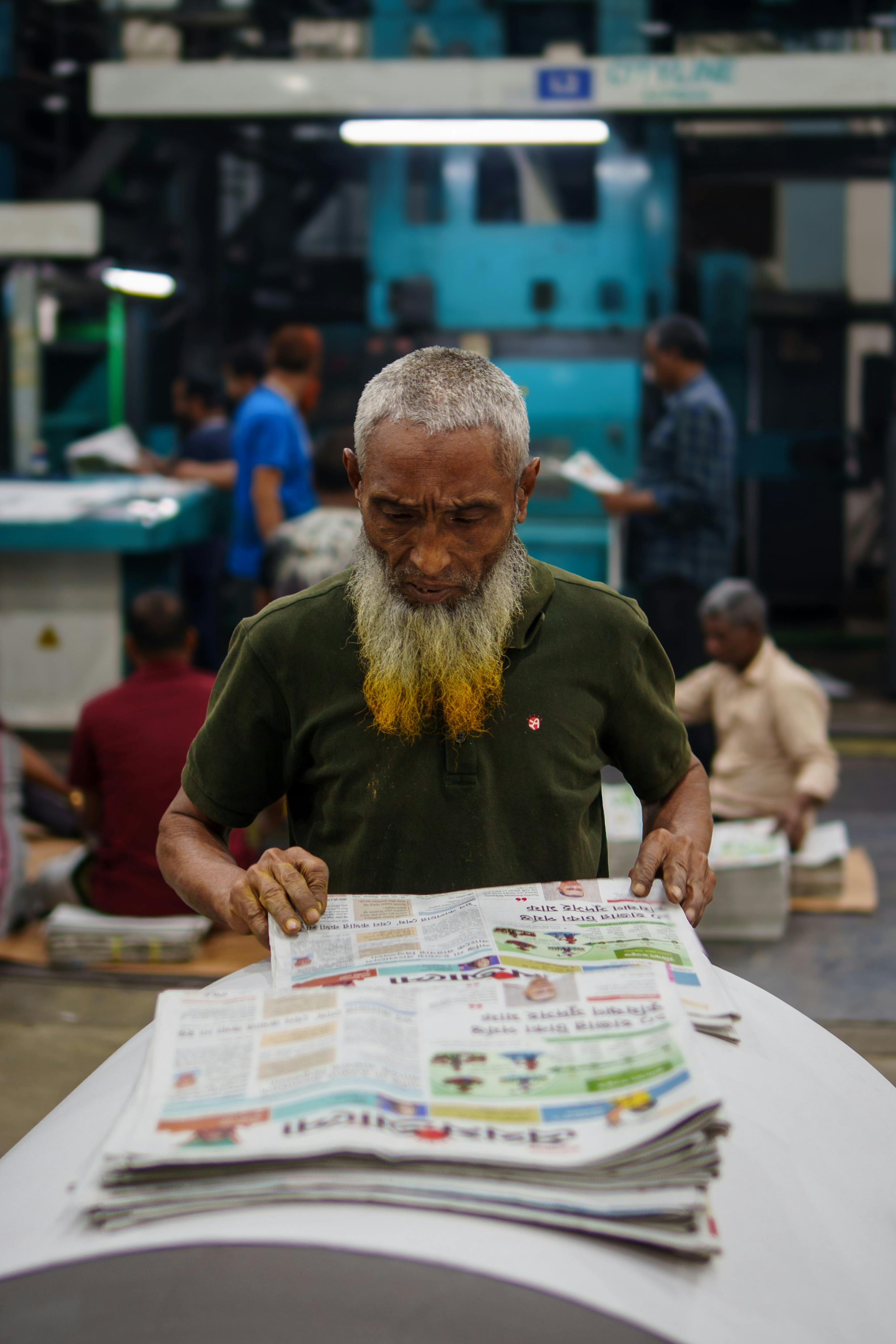 An elderly man with a yellow beard reads a newspaper in a bustling printing press in Bangladesh.