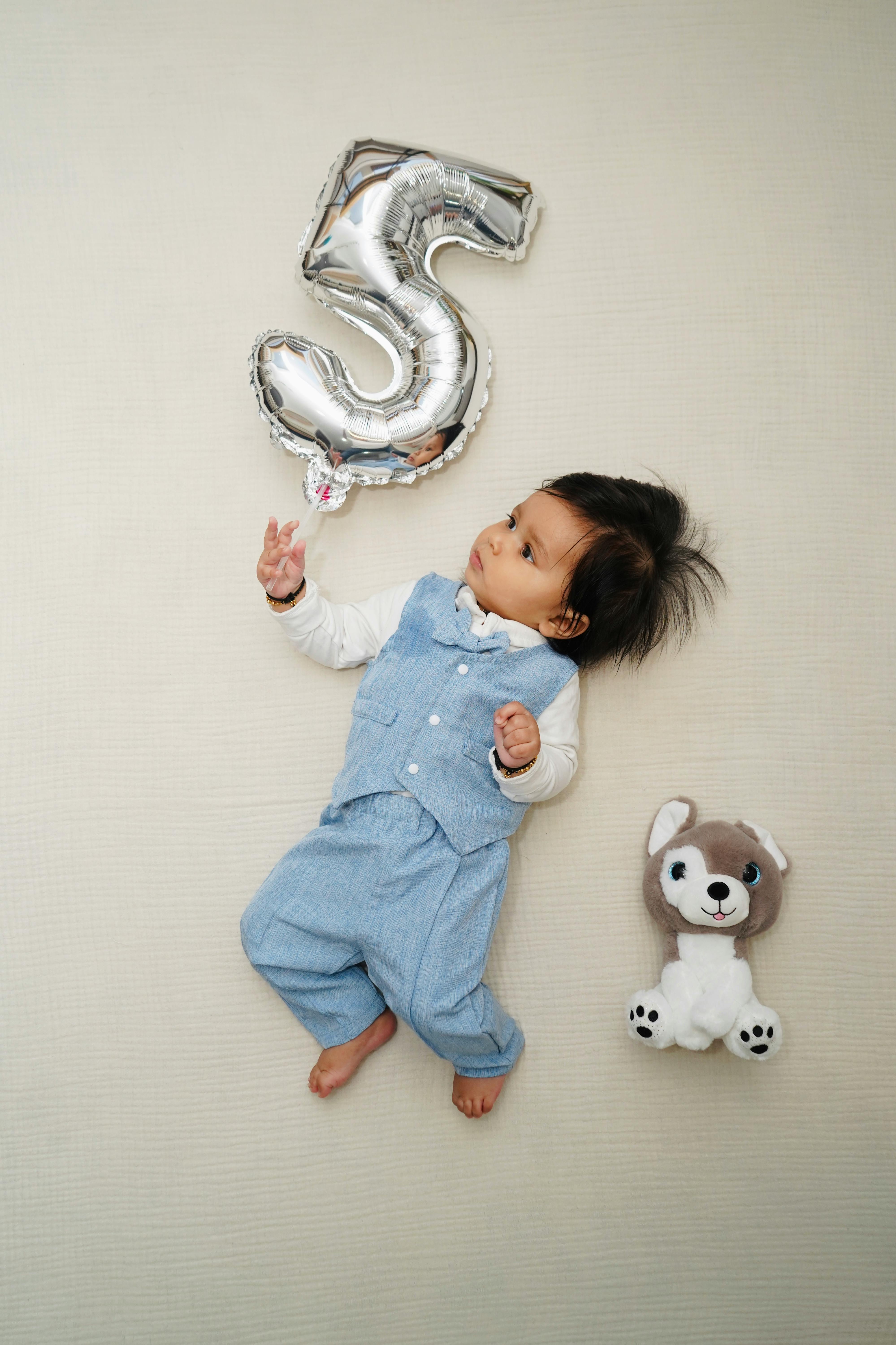 Cute baby holding a silver number 3 balloon with a stuffed toy on a light background.