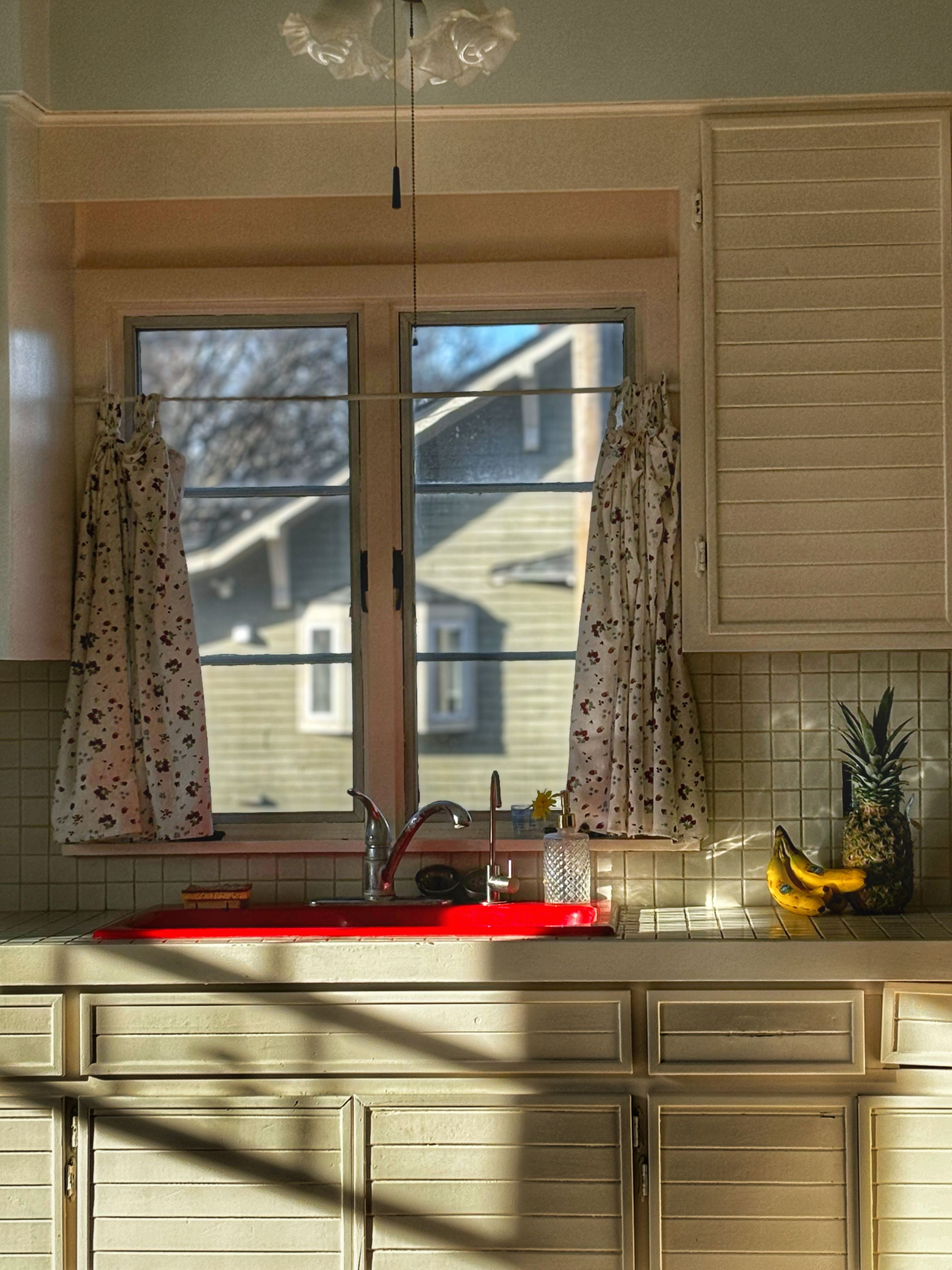 A sunlit kitchen window with a sink, curtains, and fruit arrangement, creating a warm and inviting atmosphere.