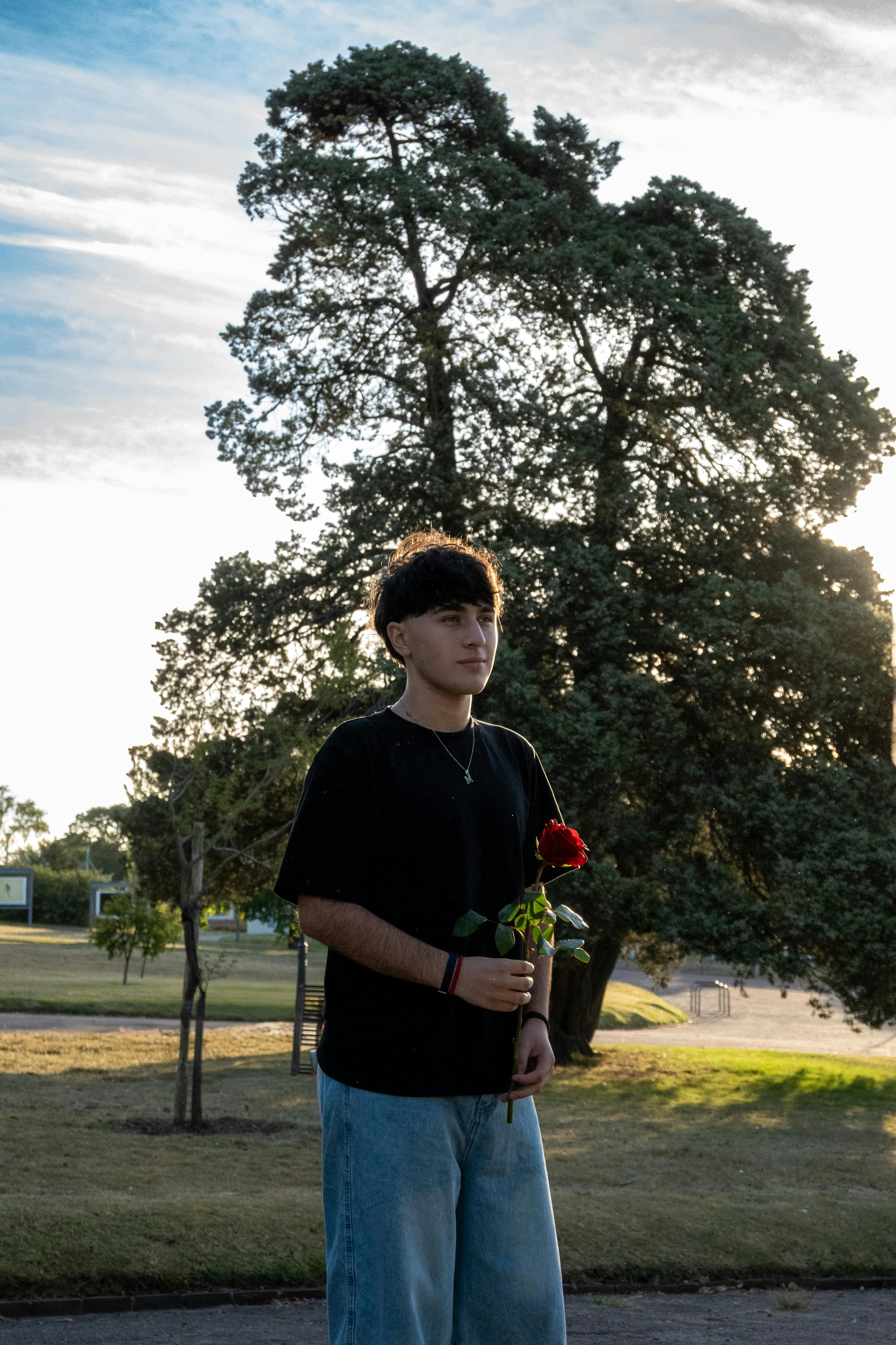 A young adult stands in a park holding a red rose, with trees and a clear sky in Montevideo, Uruguay.