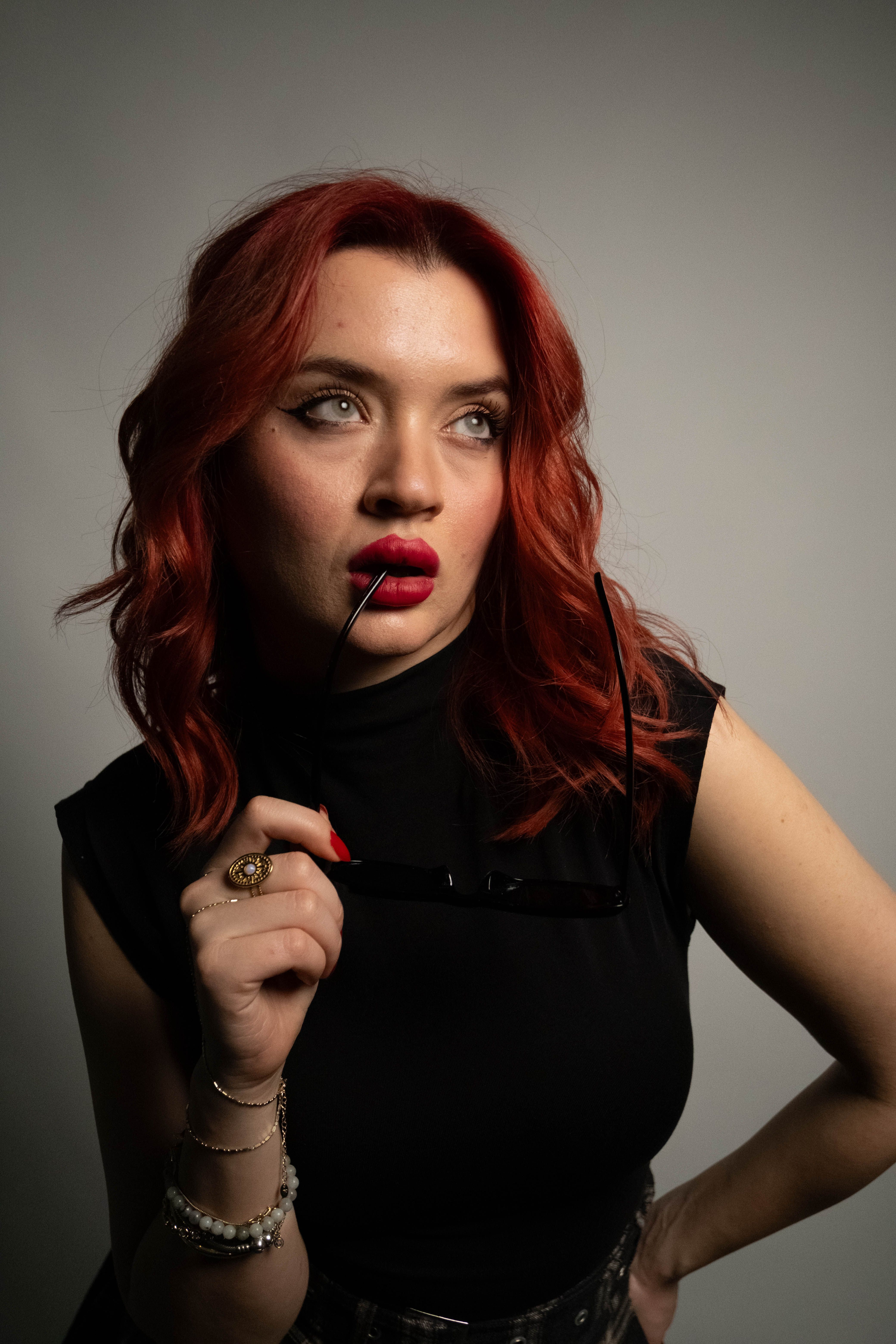 Stylish woman with red hair, deep red lipstick, posing indoors with sunglasses in hand.