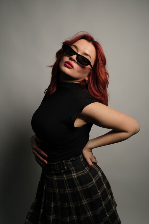 Free Fashionable woman with red hair and sunglasses posing confidently in a studio setting. Stock Photo