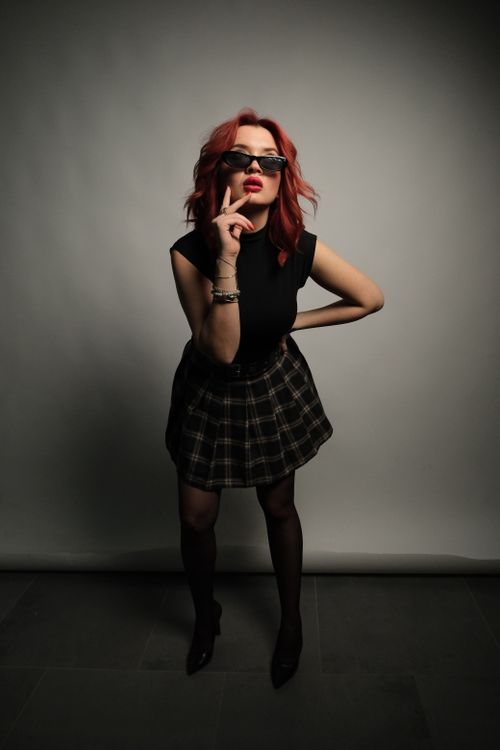 Free Red-haired woman in black top and plaid skirt poses confidently indoors. Stock Photo