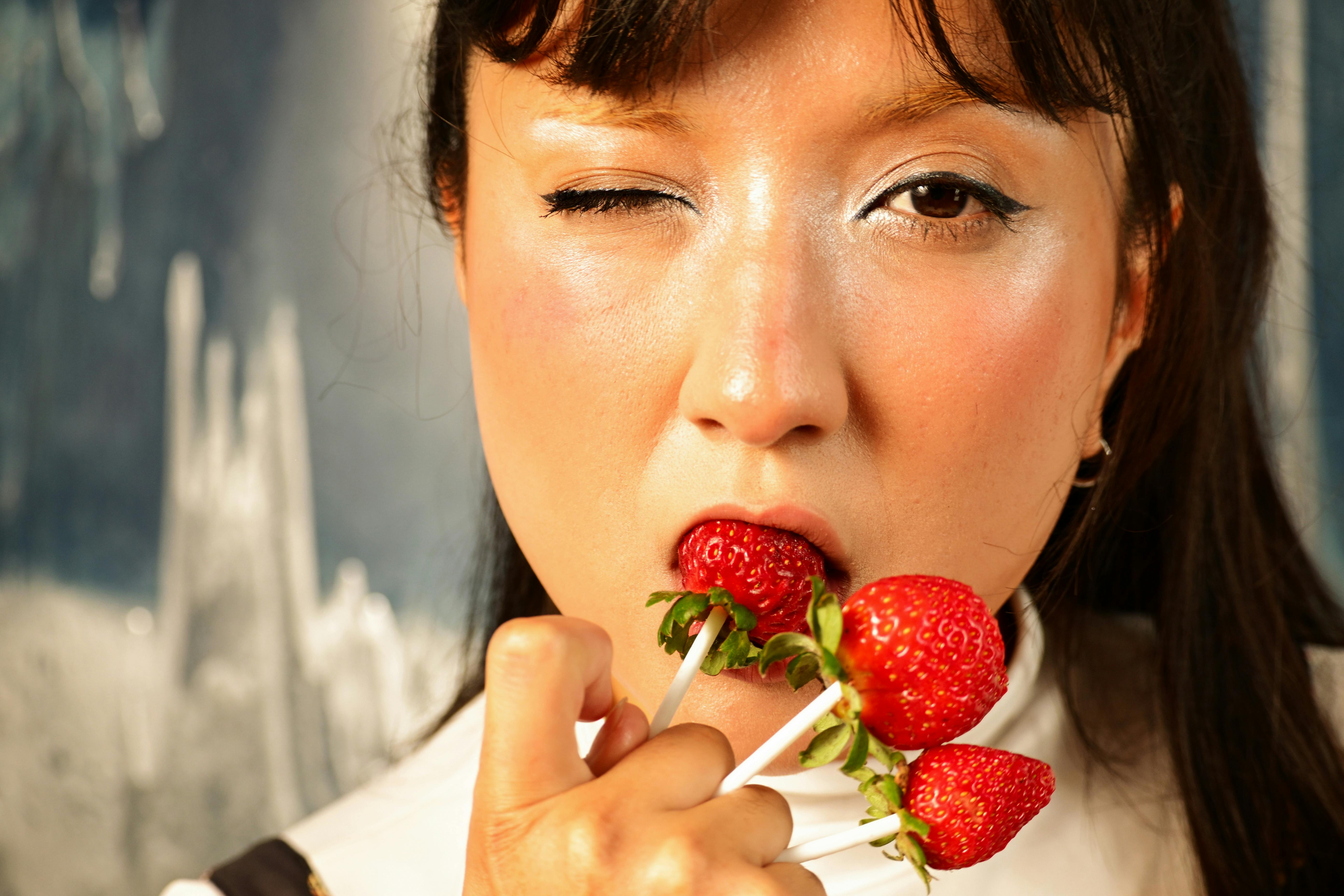 Gratis Nærbilde av en kvinne som blunker mens hun holder jordbær på pinner, noe som fremhever friskhet og moro. Arkivbilde