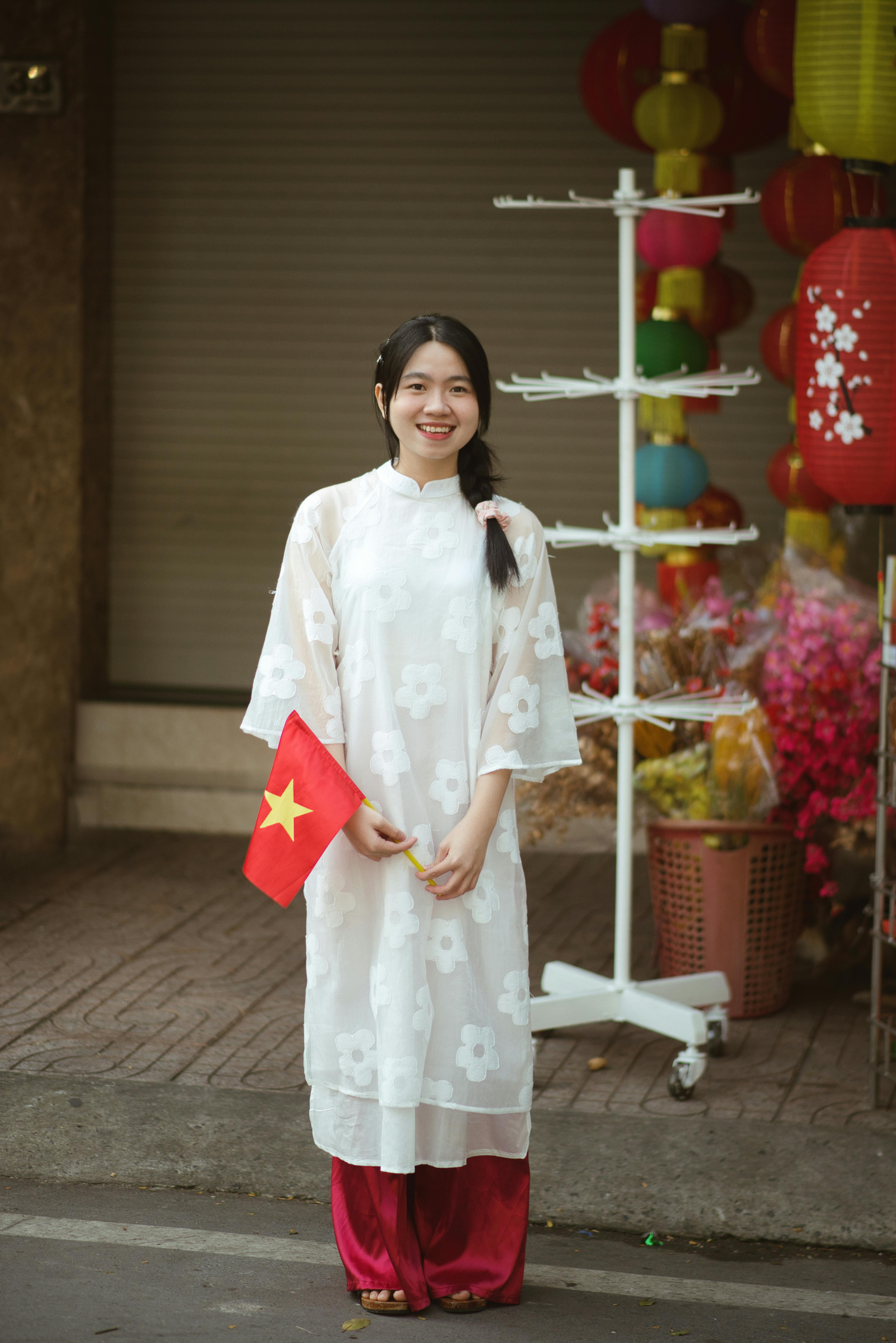 Smiling young woman in traditional white ao dai holding Vietnam flag outdoors.