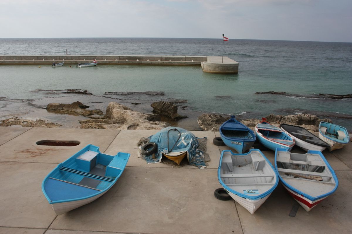 Small blue and white wooden fishing boats pulled up on the stone quay of Byblos's old harbour, beside a low breakwater and a flagged pier.