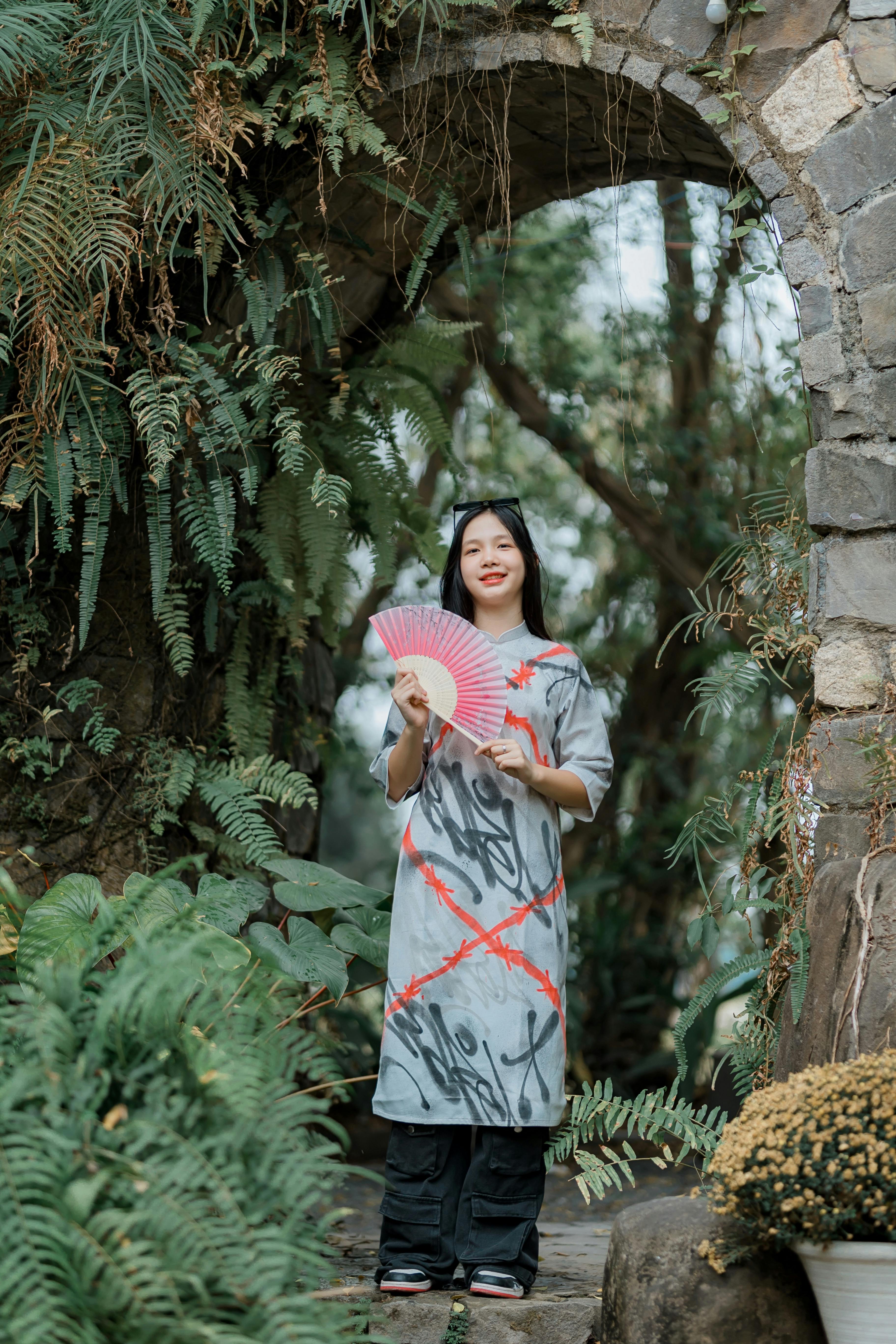 De franc Una dona que sosté un ventall rosa es troba en un exuberant jardí amb arc, envoltada de vegetació. Foto d'estoc