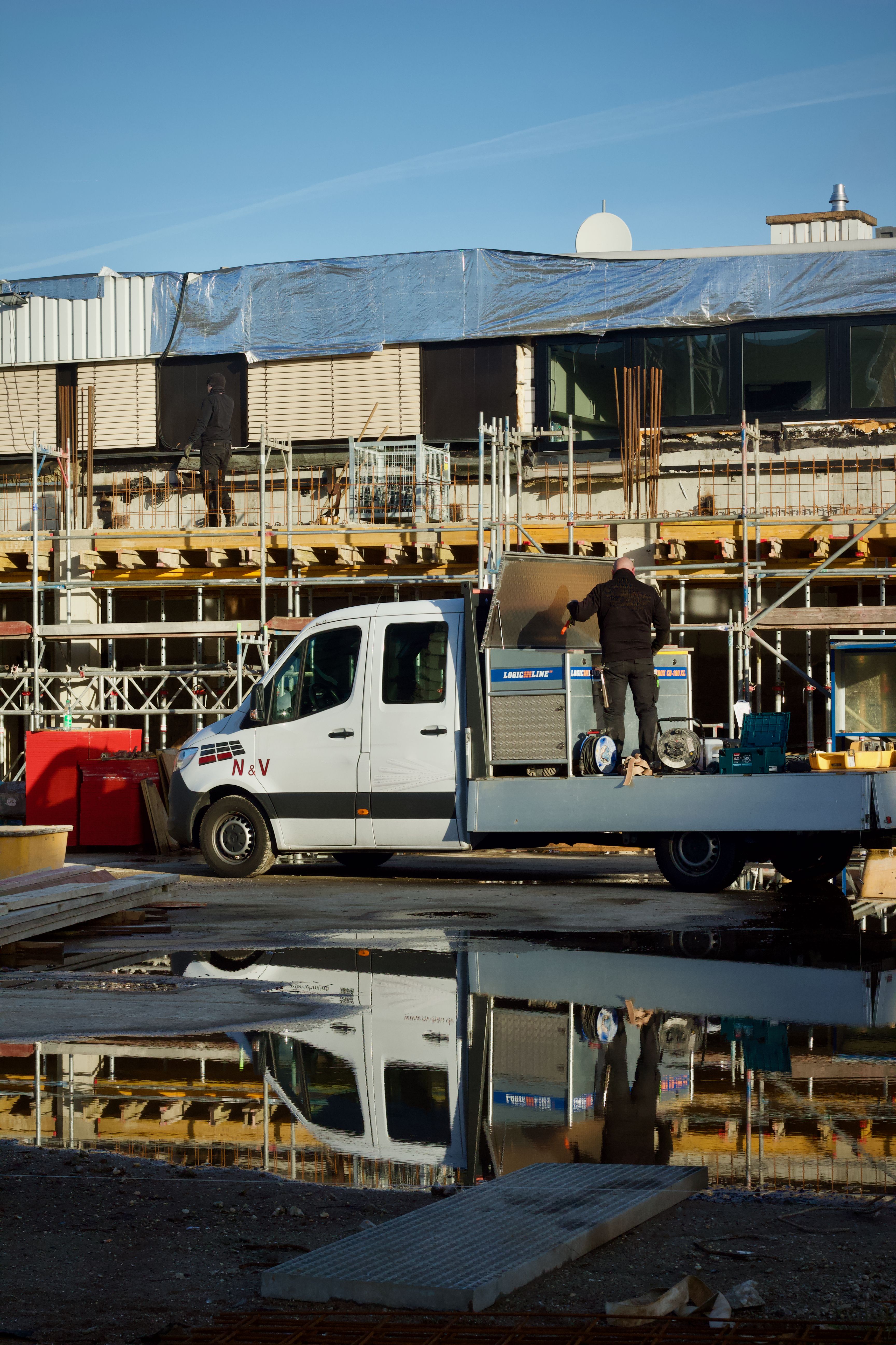 Construction worker handling equipment at building site, reflecting in water.