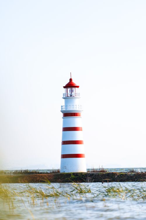Free A striking red and white lighthouse standing tall by the sea under a clear sky. Stock Photo