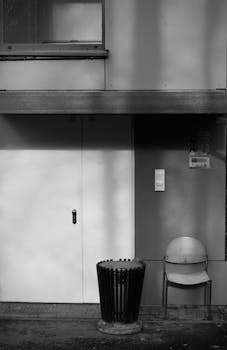 Black and white urban scene featuring a chair and trash bin beside a modern building entrance.