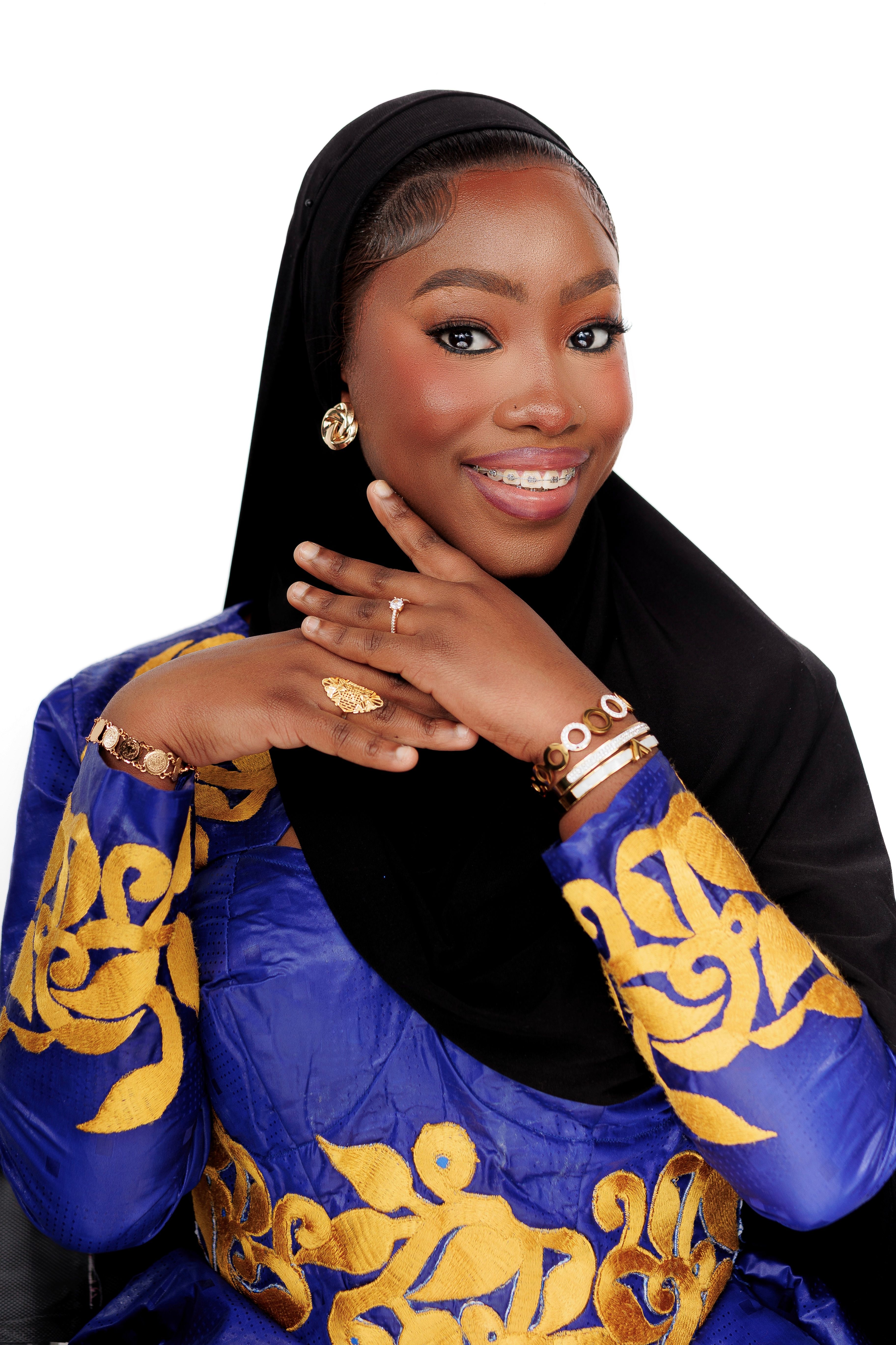 Smiling woman wearing a blue and gold dress with jewelry and a black hijab, posing elegantly.