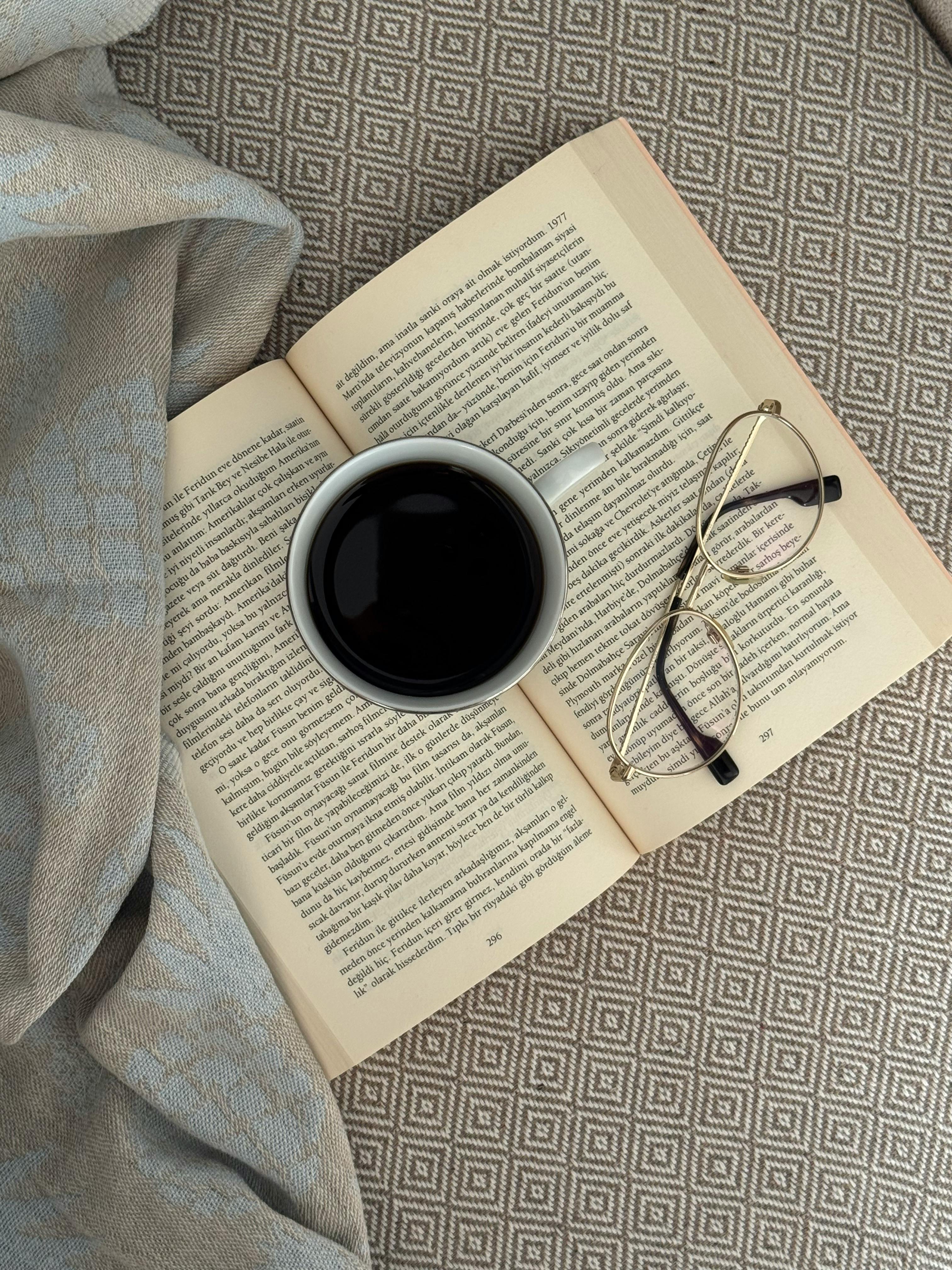 Gratis En hyggelig opsætning med en åben bog, sort kaffe og briller på et mønstret stof. Lagerfoto