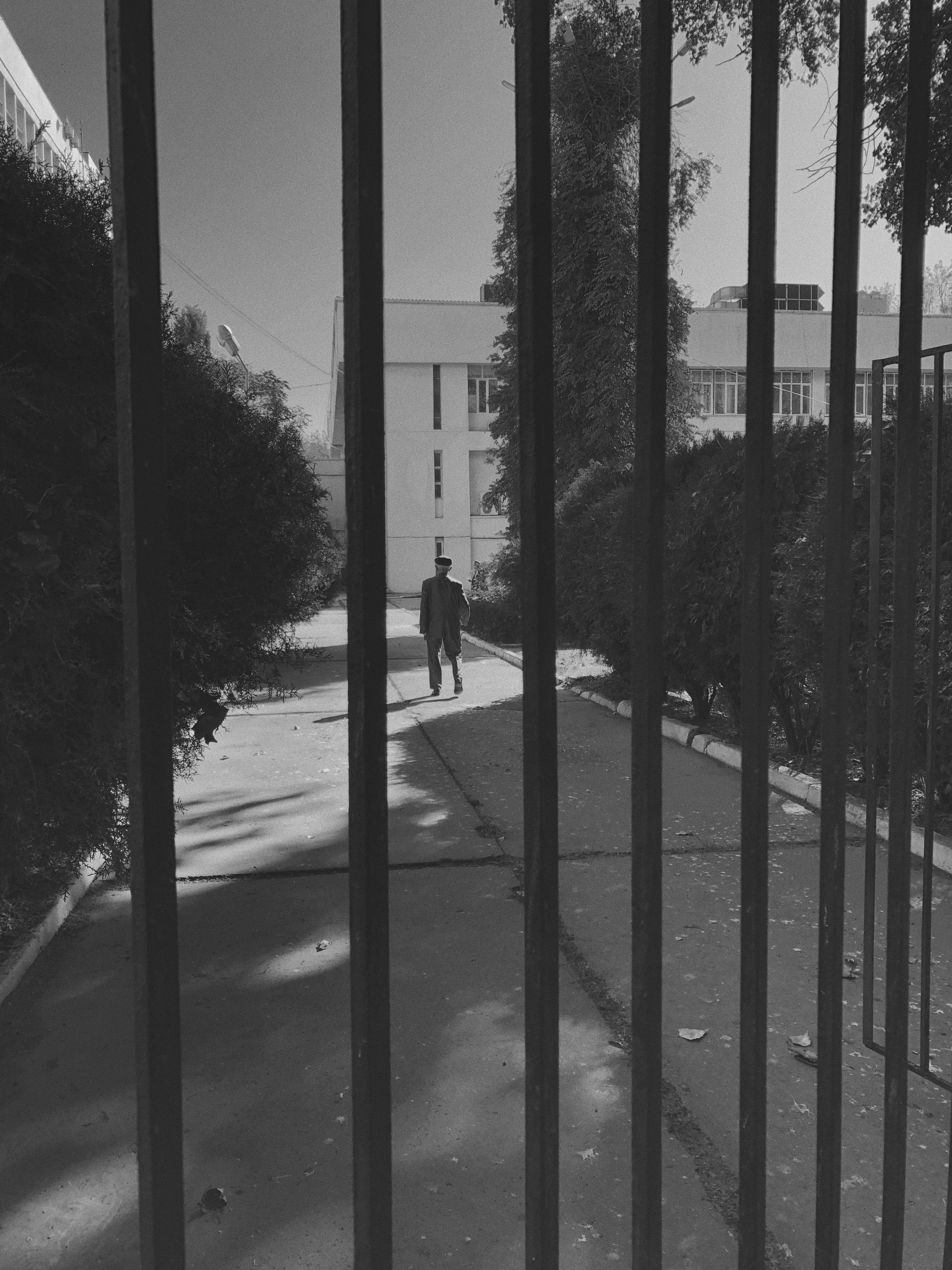 Gratuit Dans un décor urbain monochrome, une personne marche seule derrière des barreaux métalliques, évoquant l'isolement. Photos