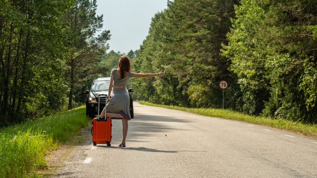 A woman hitchhiking with an orange suitcase on a rural road in Estonia during summer.