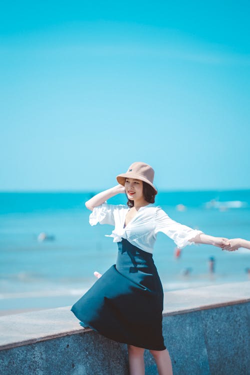 Женщина в белой рубашке с длинным рукавом и черной юбке в белой шляпе стоит на берегу моря
