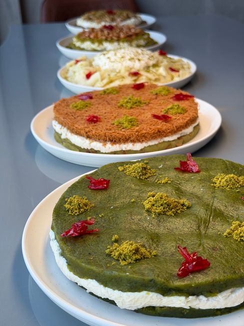 A sliced layer cake showing dark chocolate cake layers separated by a golden, crunchy pistachio-kunafa filling, coated in glossy milk chocolate ganache and topped with chopped pistachios.