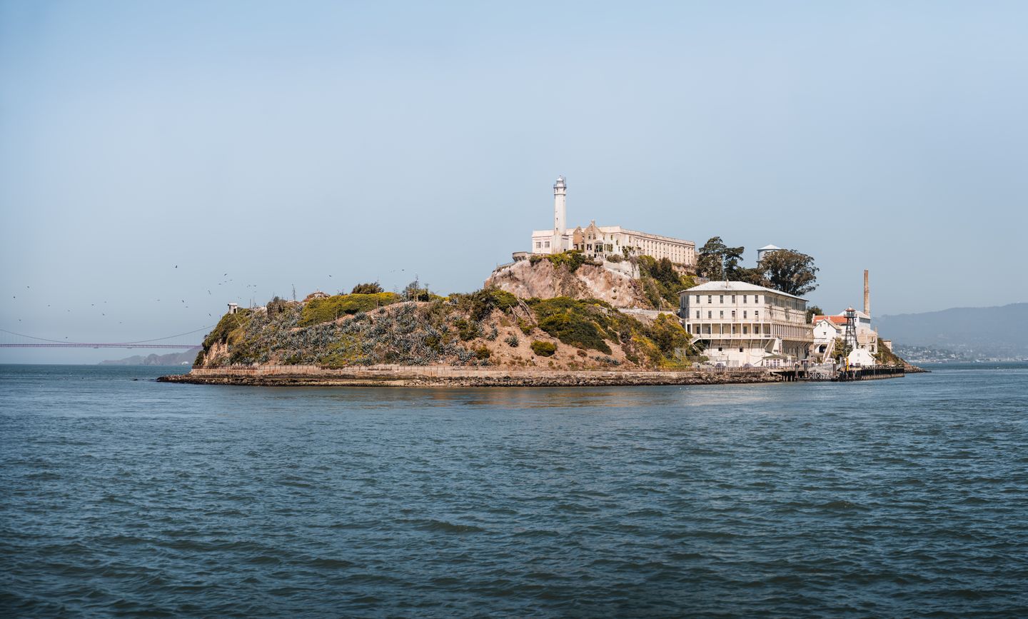 Alcatraz Island with its lighthouse and prison buildings seen from the bay under clear skies, with the Golden Gate Bridge visible in the distance