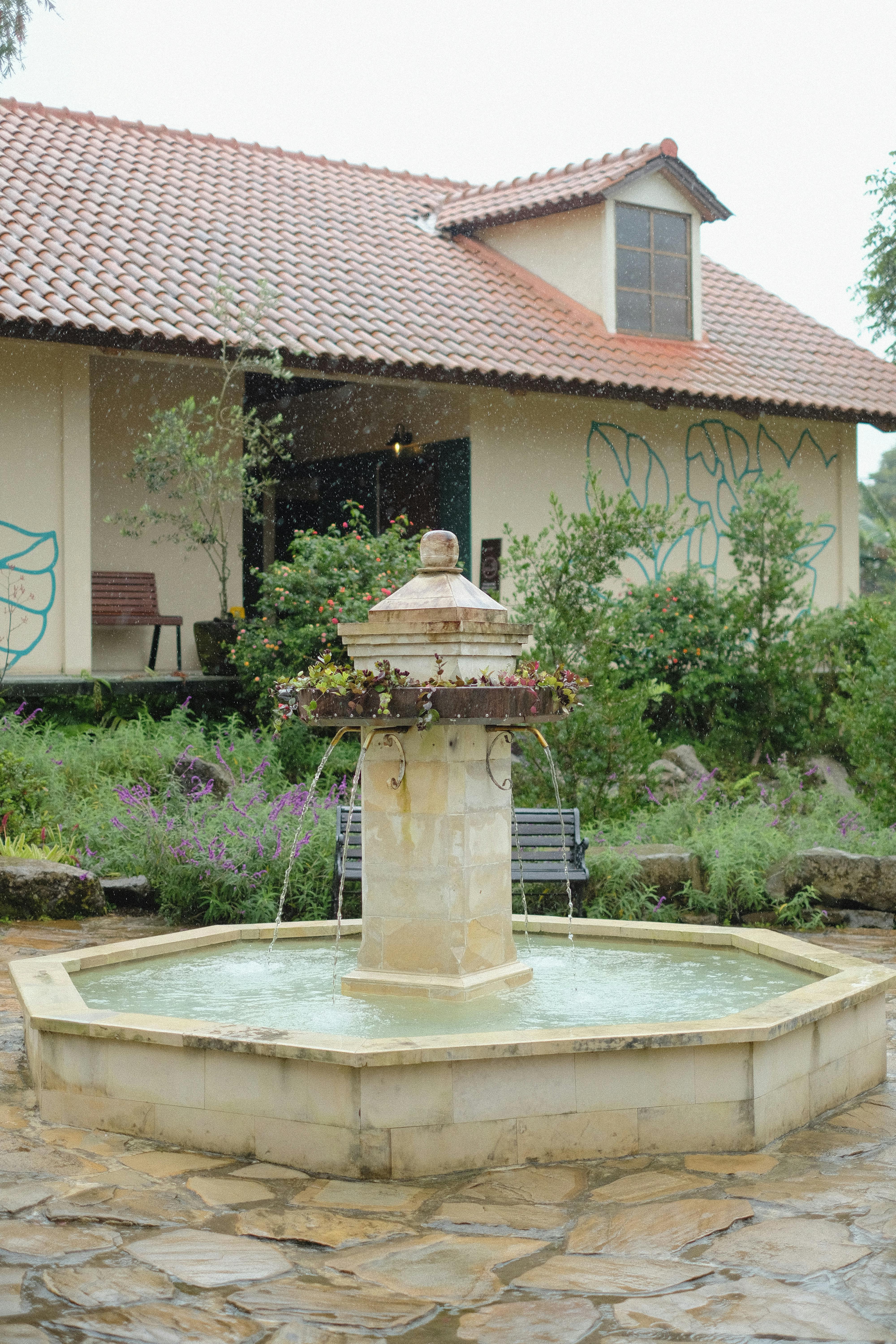 бесплатная Безмятежный сад с декоративным фонтаном, окруженный зеленью, и живописным домиком на заднем плане. Стоковое фото