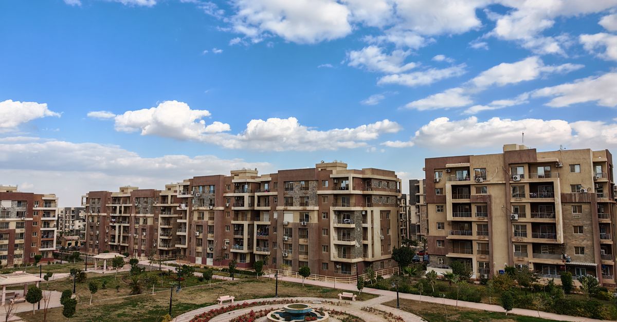Aerial view of a modern residential complex with a central fountain under a blue sky and clouds.