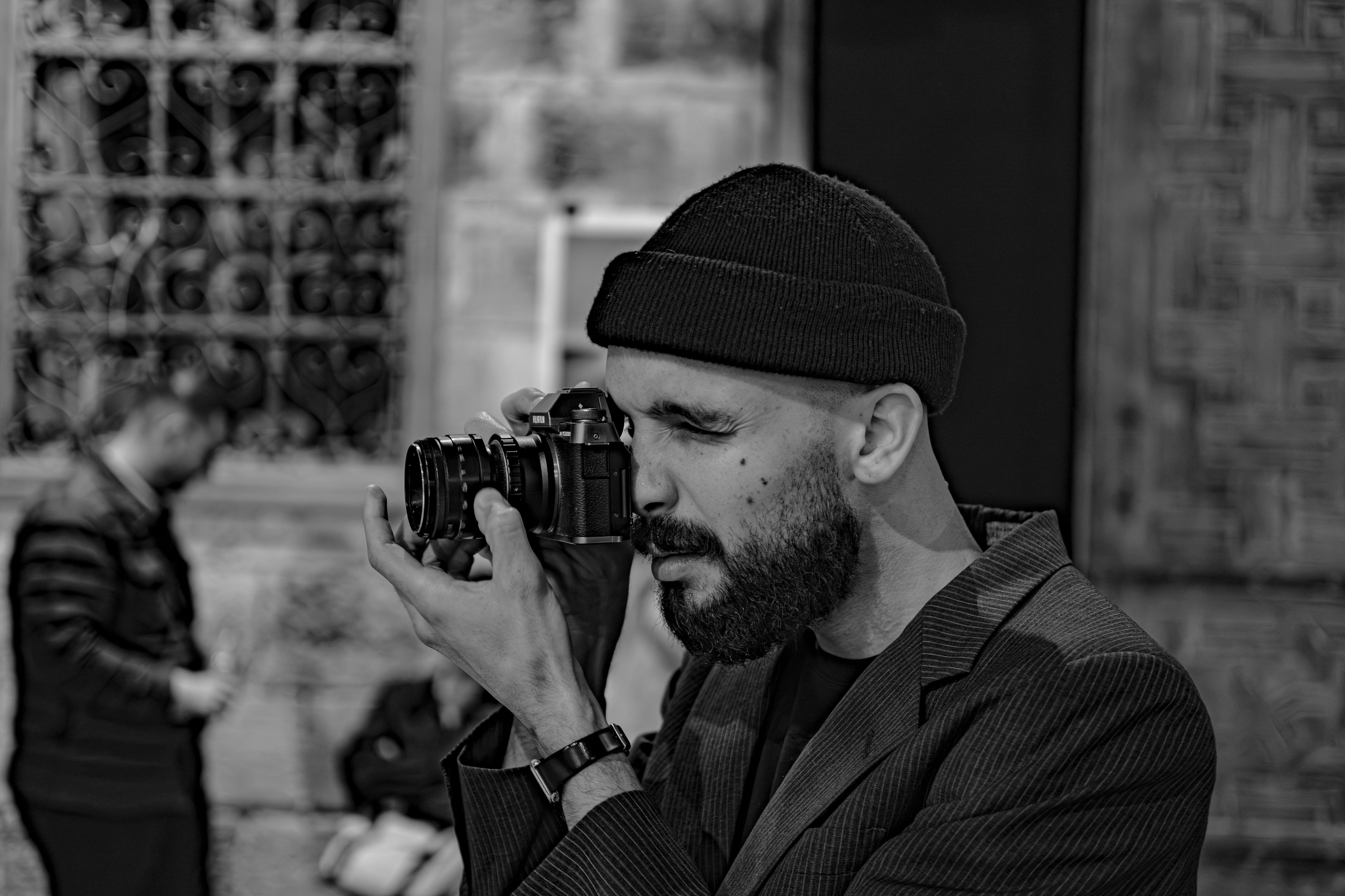 A man with a beard and beanie takes a photo with a DSLR on a city street in monochrome.