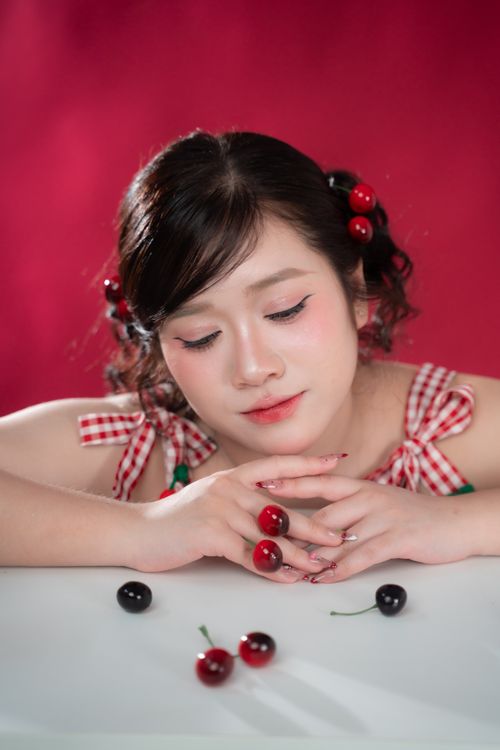 Free Young woman with cherry-themed hairstyle and clothing poses thoughtfully against a vibrant red backdrop. Stock Photo