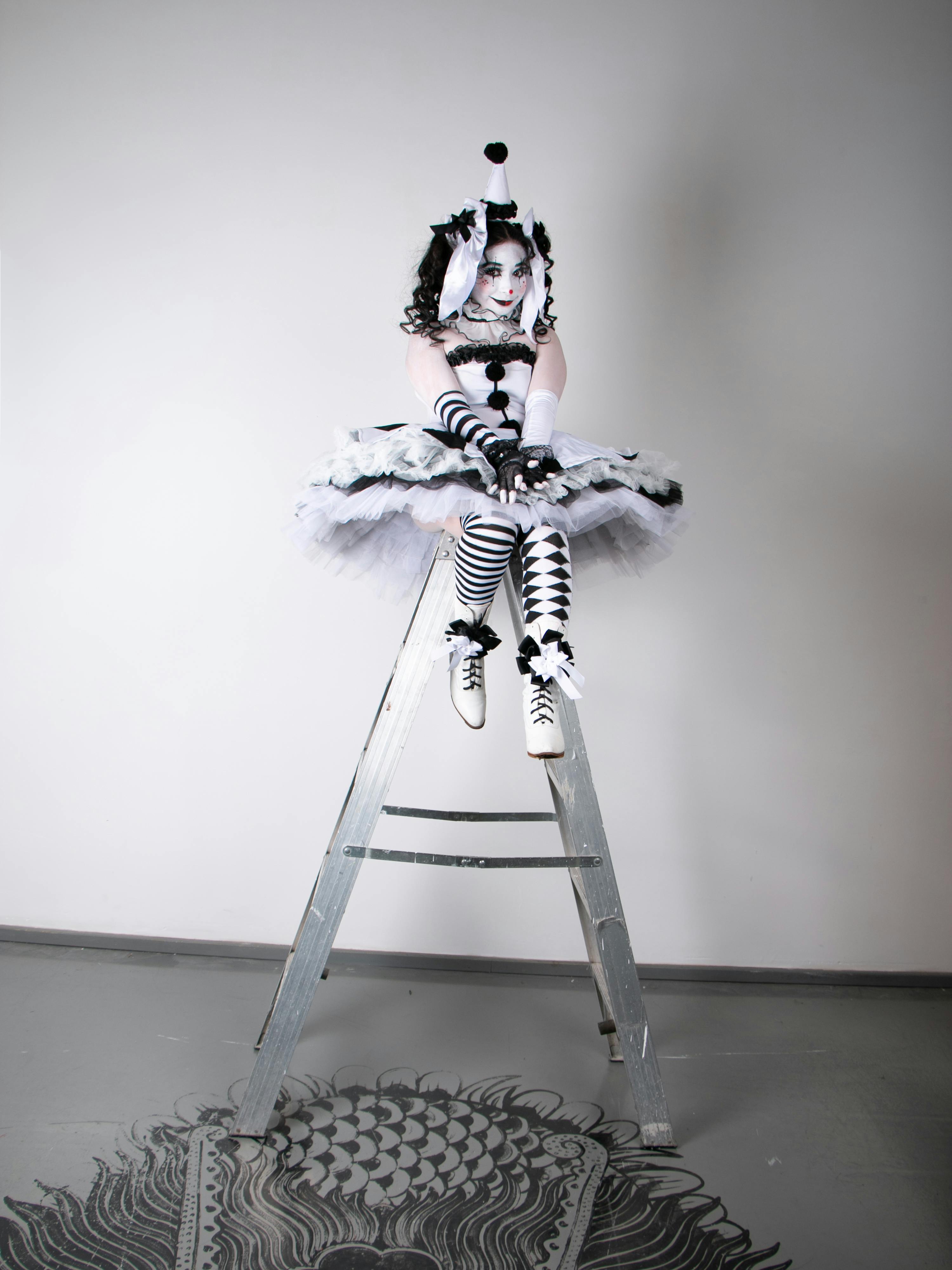 Gratuit O persoană într-un costum de clovn alb-negru stă pe o scară într-o cameră minimalistă. Fotografie de stoc