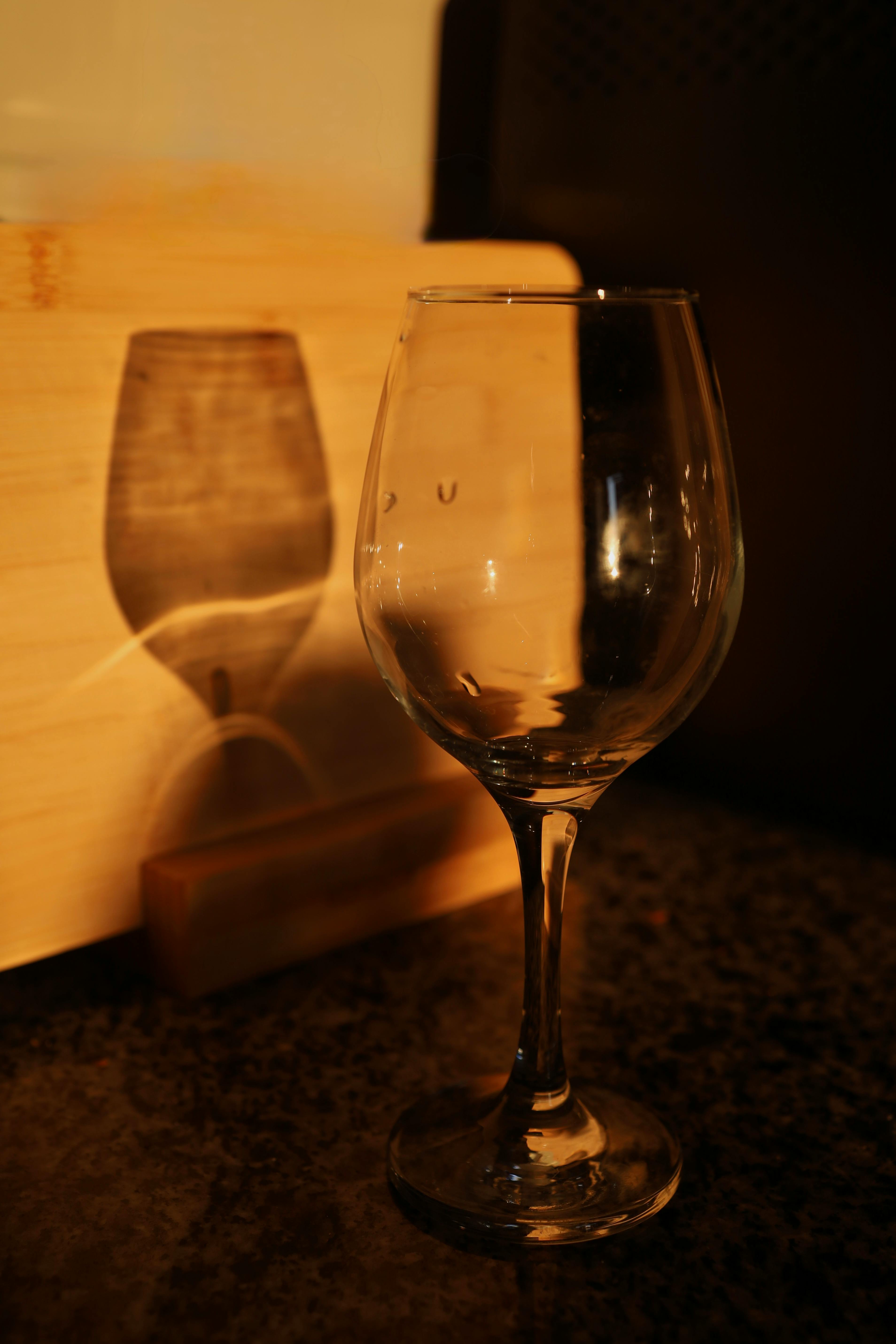 Free A clear wine glass with its shadow cast on a wooden surface in warm lighting. Stock Photo