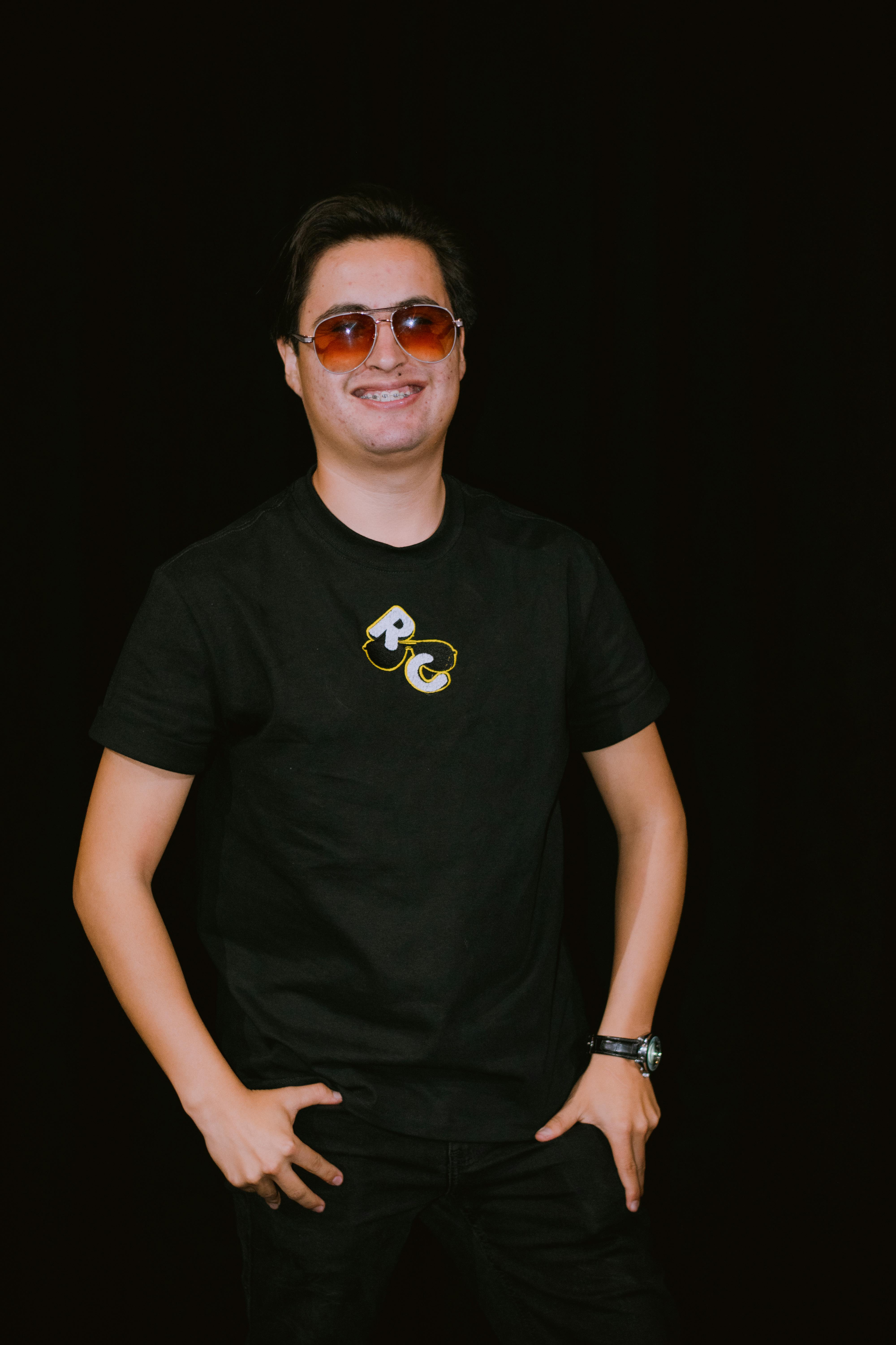 Cheerful man wearing sunglasses and black t-shirt poses confidently against a solid black backdrop.