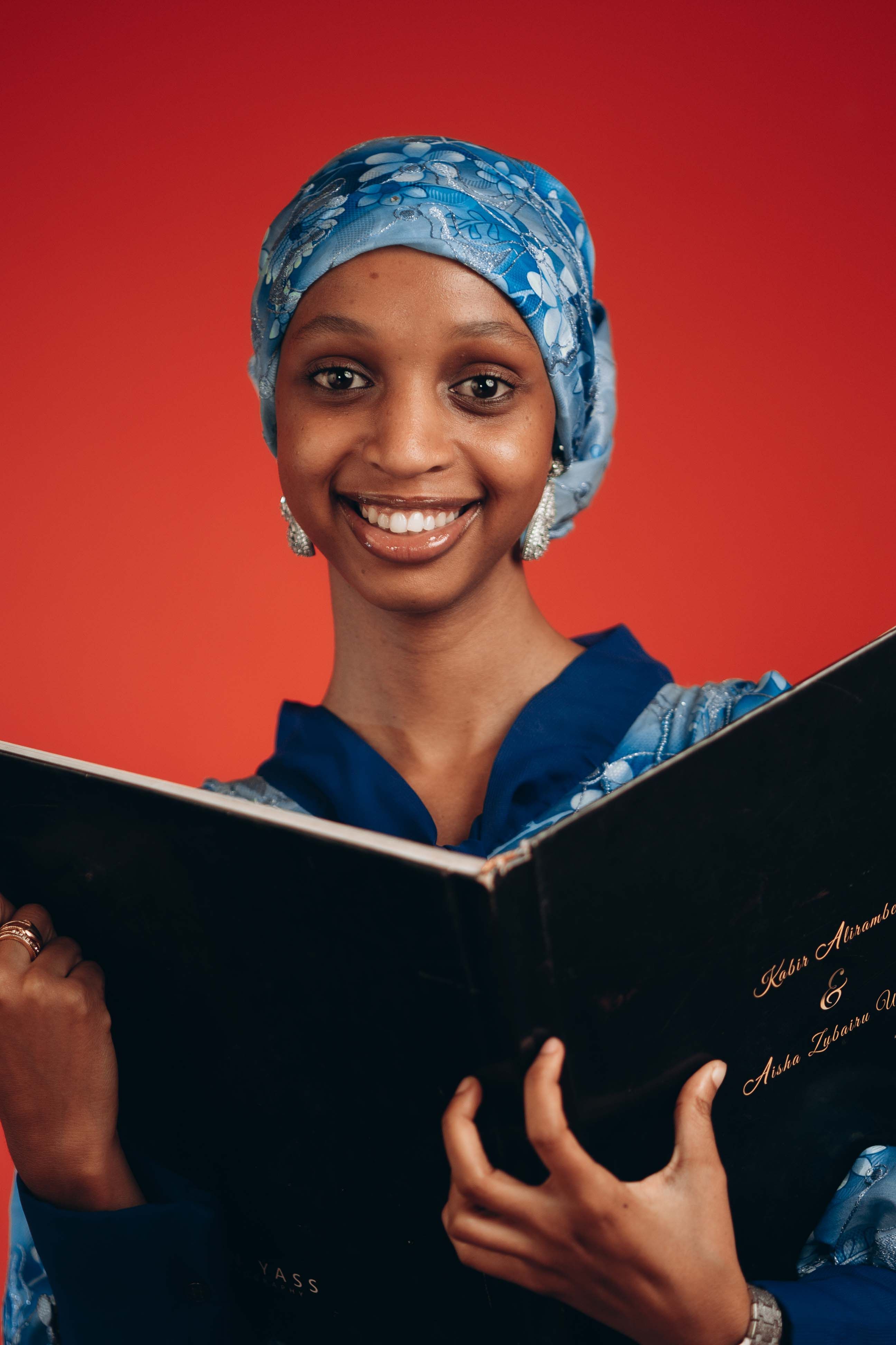 A woman wearing a blue headscarf and dress smiles while reading a book indoors against a red background.