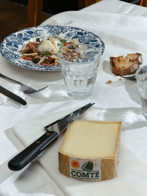 A wedge of aged Comté cheese showing its dense ivory interior and small scattered holes, next to a whole cloth-bound wheel with its natural brown rind.