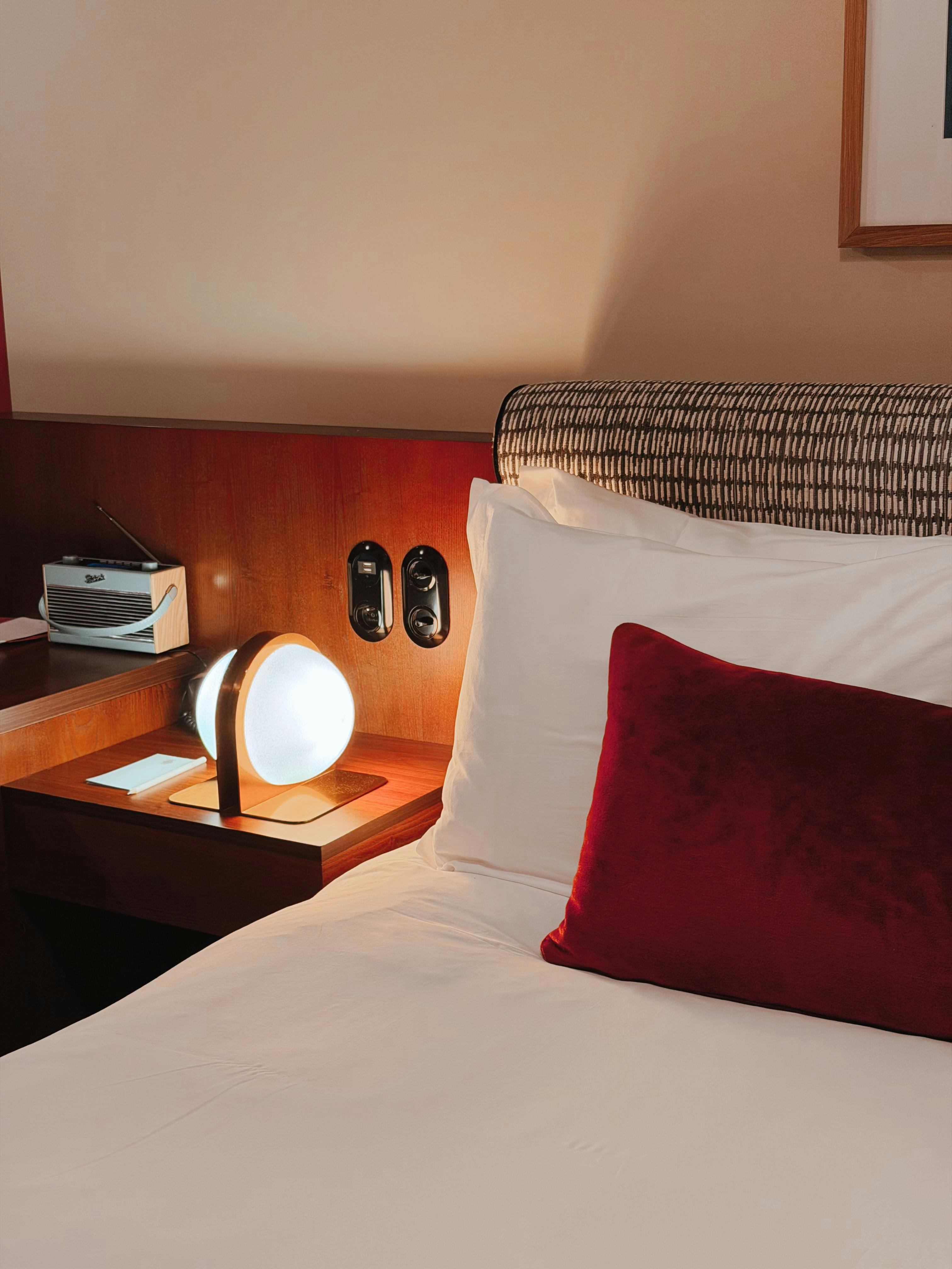 Elegant hotel room interior with bedside lamp and plush red pillow on white bedding.