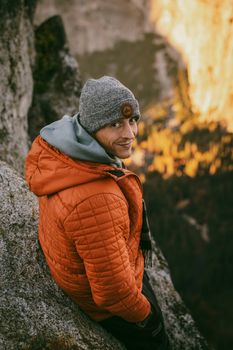 A man in a cozy orange jacket and gray beanie enjoys a mountain view at sunrise.