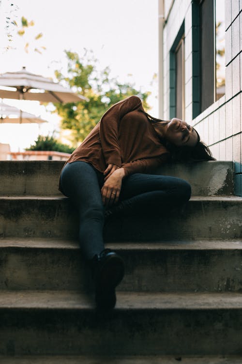 Woman in Brown Long Sleeved Top and Black Pants Sitting While Sleeping on Gray Concrete Stairs