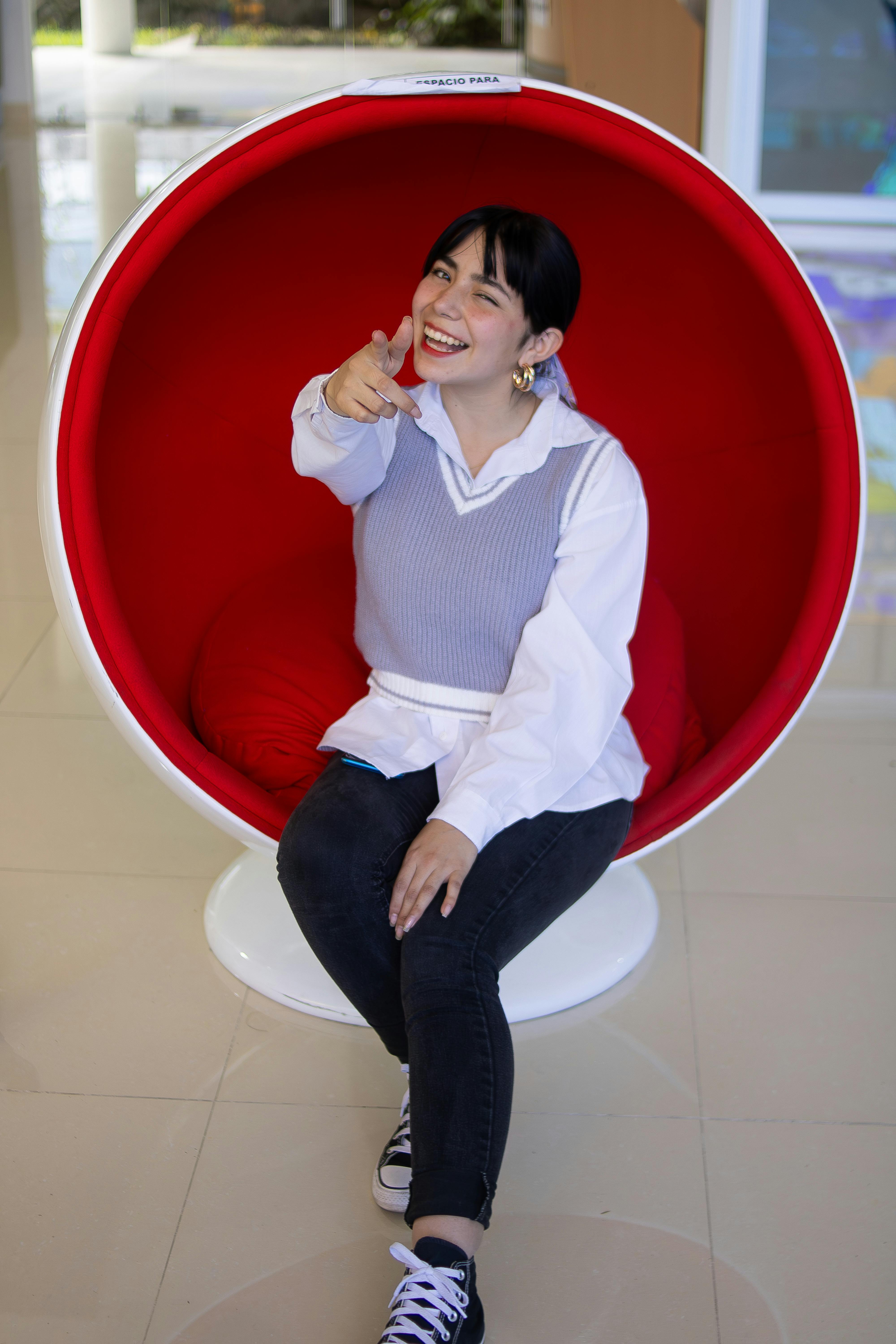 無料 赤いポッドチェアに座り、室内で指さしながら微笑んでいる幸せそうな女性。 写真素材