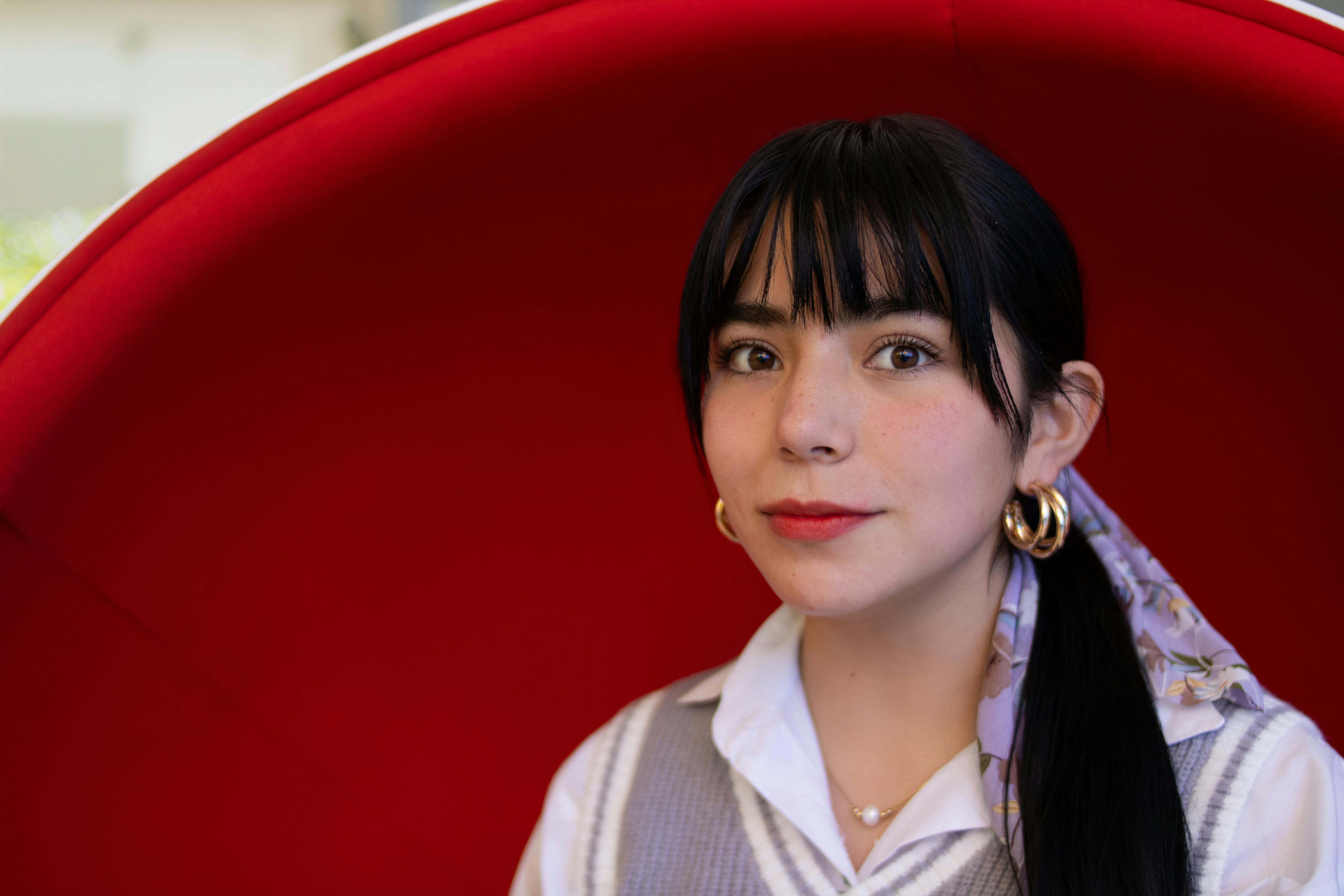 Free Portrait of a young woman seated in a modern red chair, exuding elegance with subtle makeup and accessories. Stock Photo