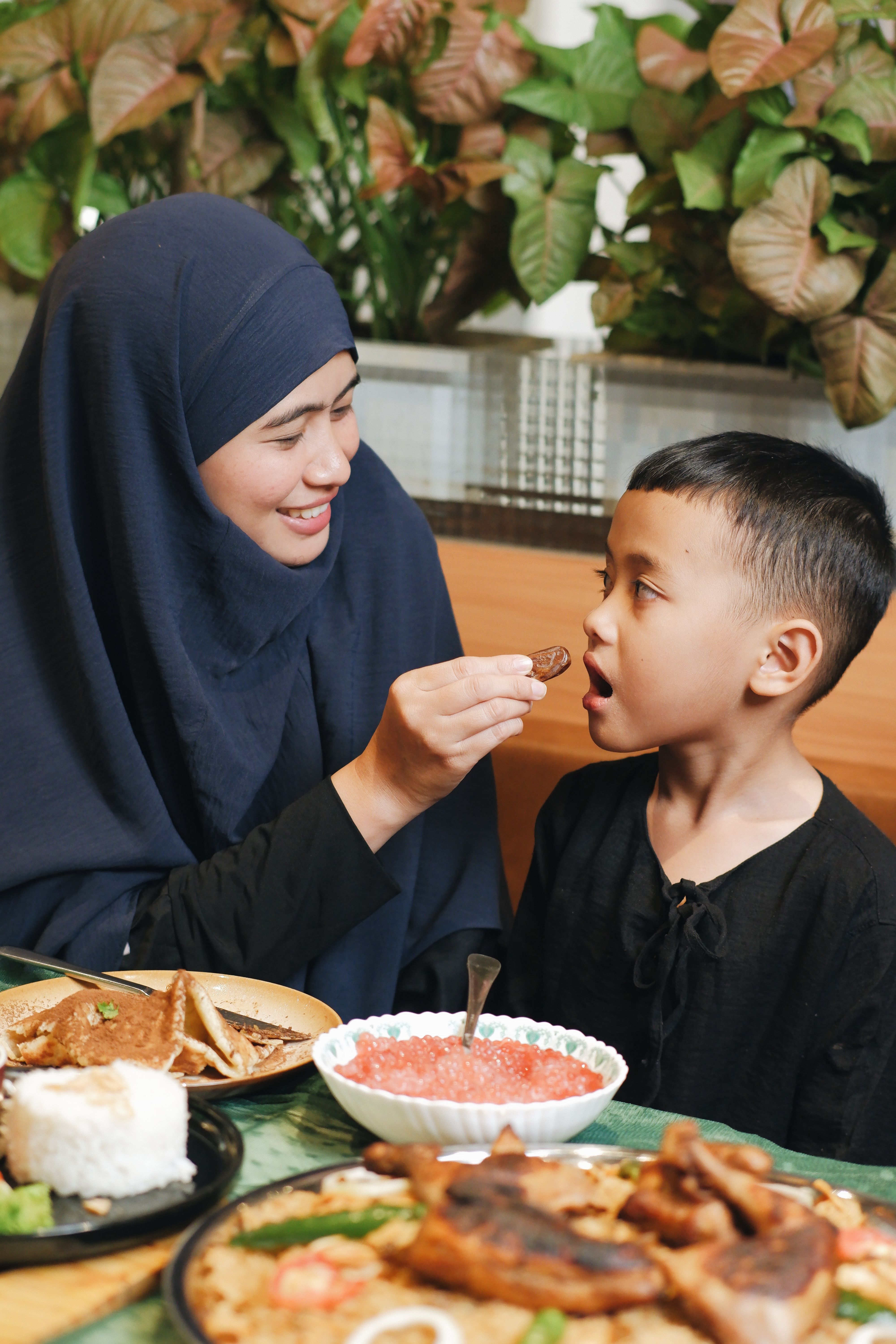 Muslim mother in hijab feeding her son during a meal in a cozy setting, capturing a warm family moment.
