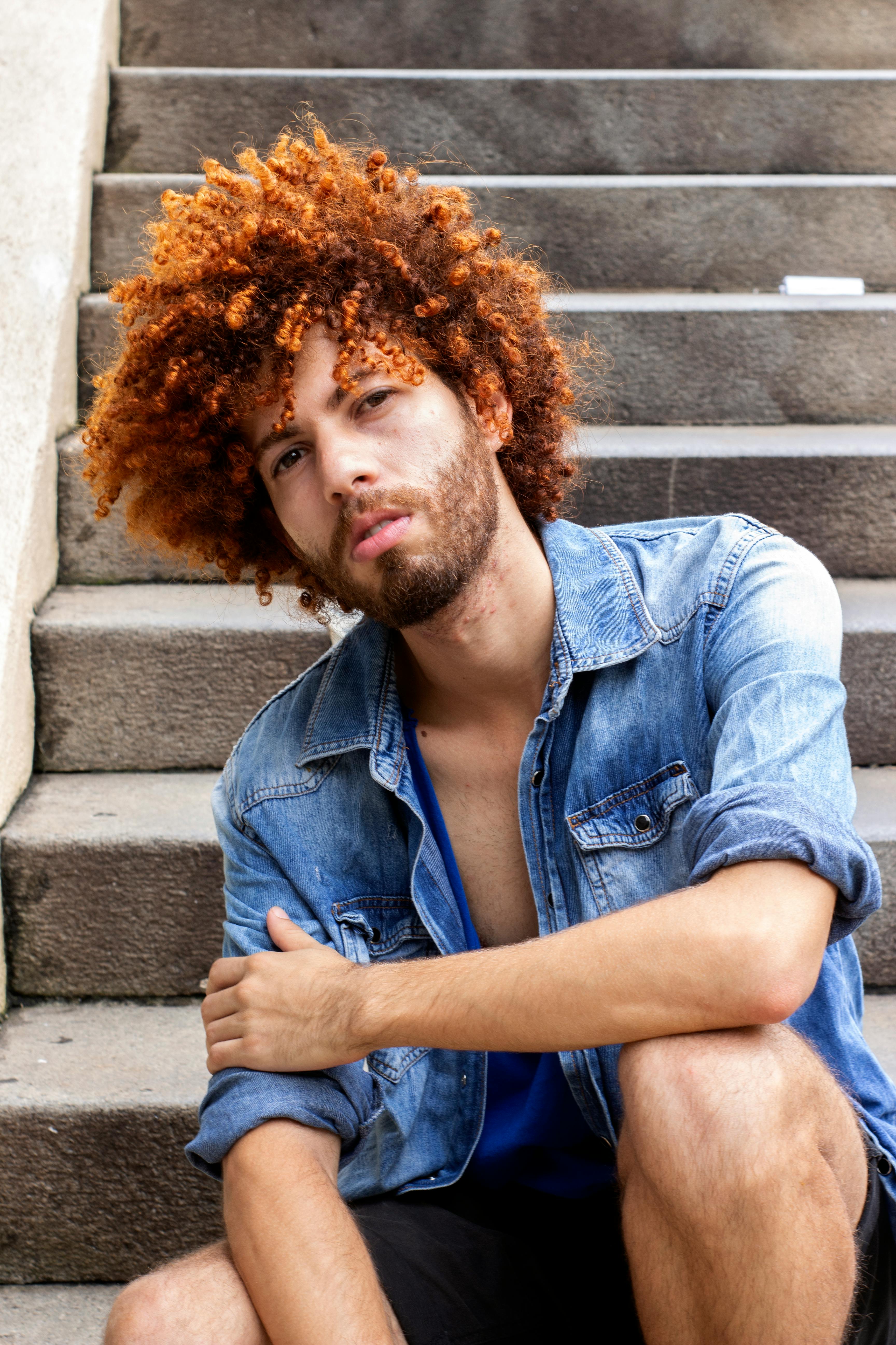 一个时尚的年轻人，留着卷发，穿着牛仔夹克，表情严肃地坐在混凝土台阶上