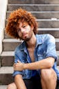 一个时尚的年轻人，留着卷发，穿着牛仔夹克，表情严肃地坐在混凝土台阶上