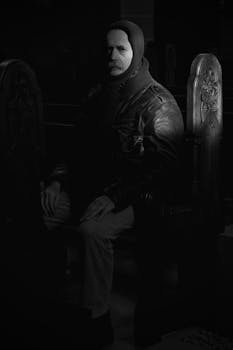 A man in a jacket sits solemnly in a dark, dimly lit room.